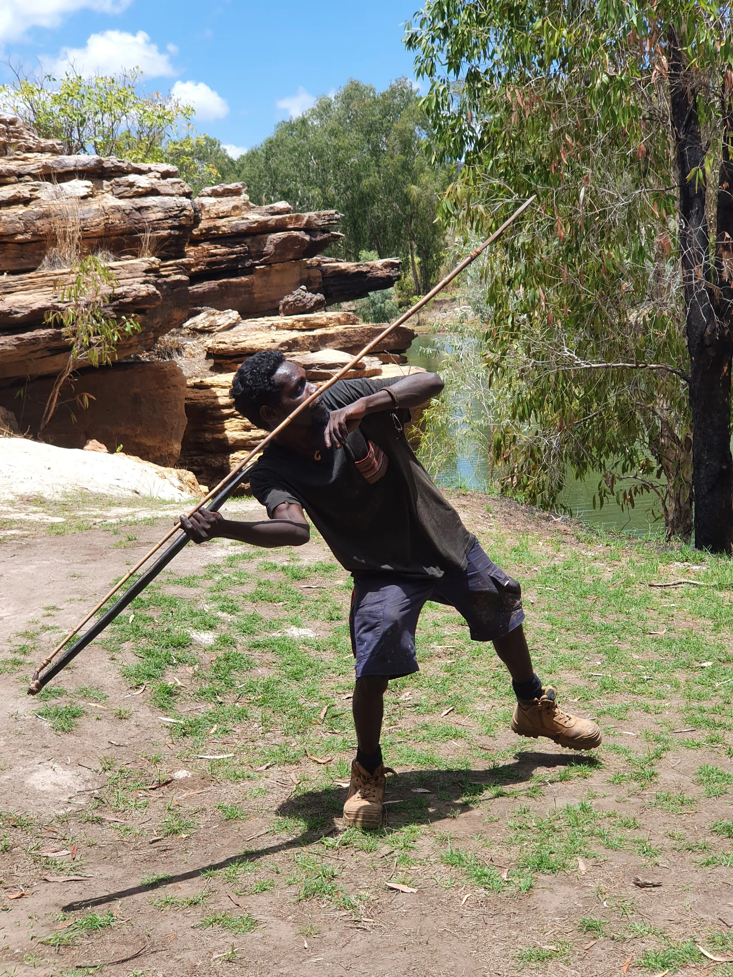 A man wearing a black shirt, gray shorts, and tan boots, is stretching outdoors on a grassy area with muddy patches. He holds a long stick or staff and is leaning back with one leg bent. There are green trees, rocks, and a small body of water in the 