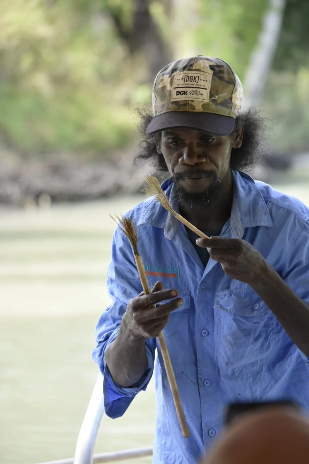A man with dark hair and a beard, wearing a camouflage cap and a light blue shirt, is holding two reeds and examining them near a body of water with trees in the background.