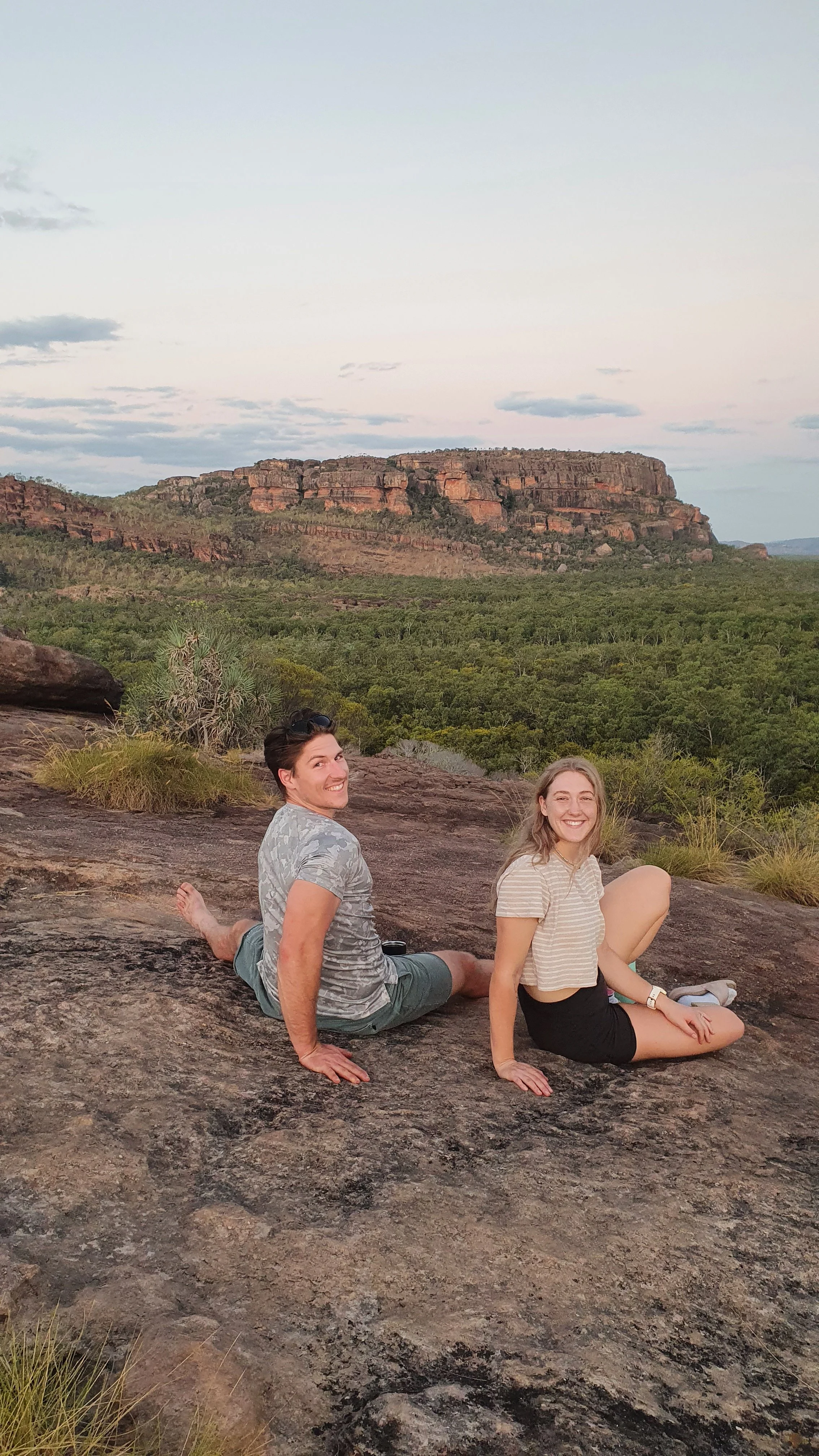 Two people sitting on a large rock, smiling at the camera with a green desert and a flat-topped mountain in the background.