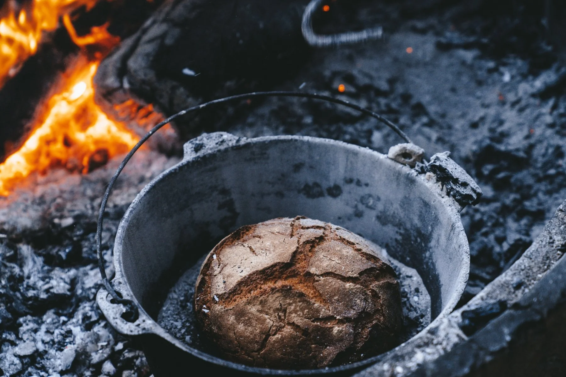A loaf of bread inside a metal bucket on a fire, with glowing coals and flames in the background. Damper - Bush Bread