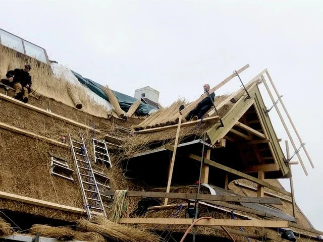 Person arbejder på tag på et byggeri med tagpaptag, lejder og stiger, under et gråt himmel, i en konstruktions- eller renoveringssituation.