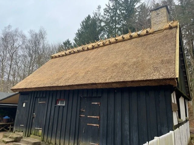 En lille, sort træhytte med stråtag, omgivet af træer i baggrunden. Tækkemand Nordsjælland Furesø Tækkeentreprise ApS