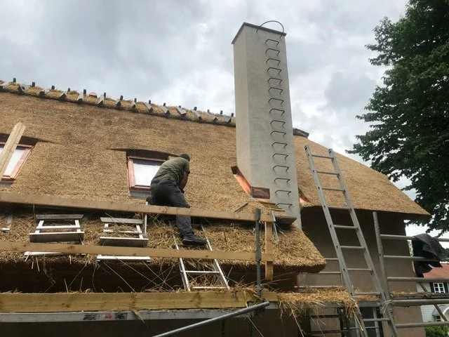 En person arbejder på taget af en hytte med stråtag, der er ved at blive renoveret, med en stor skorsten og to stiger.