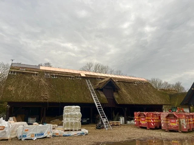 Et halmtaghus under renovering med stige, byggematerialer og affaldscontainere foran, og en overskyet himmel i baggrunden.