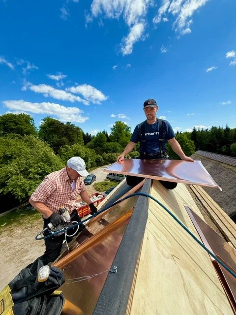 To mænd arbejder på taget af et hus under montering af kobberplader, omgivet af grønne træer og en blå himmel.