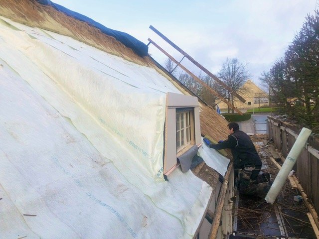 Person arbejder på taget af en bygning, som er under renovering, med isolering og plastfolie. Tækkemand Nordsjælland Furesø Tækkeentreprise ApS