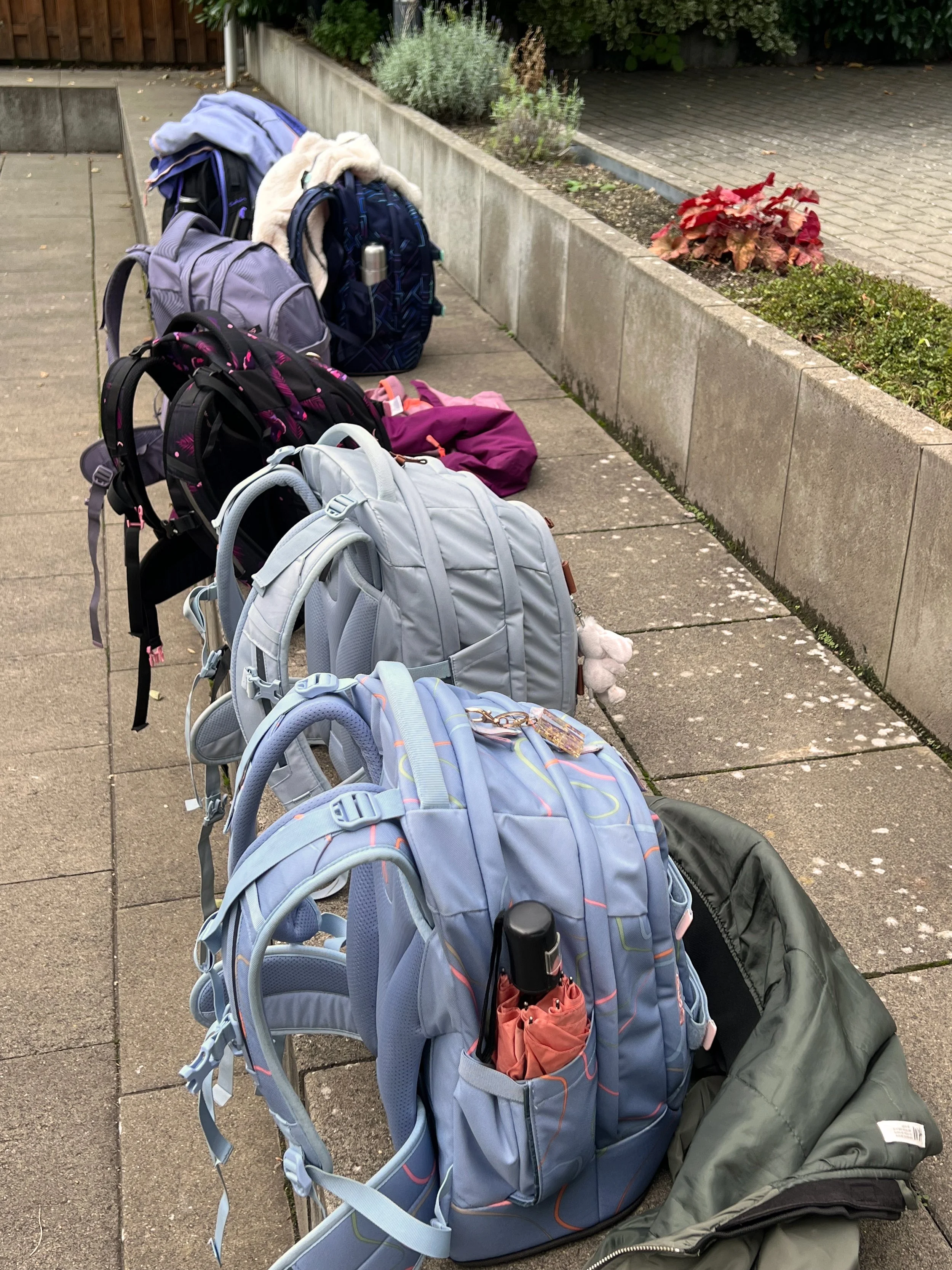 Mehrere Rucksäcke in verschiedenen Farben auf dem Gehweg, mit einigen Gegenständen wie einem Regenschirm und kleinen Anhängern. Im Hintergrund eine bepflanzte Mauer und ein gepflasterter Weg.
