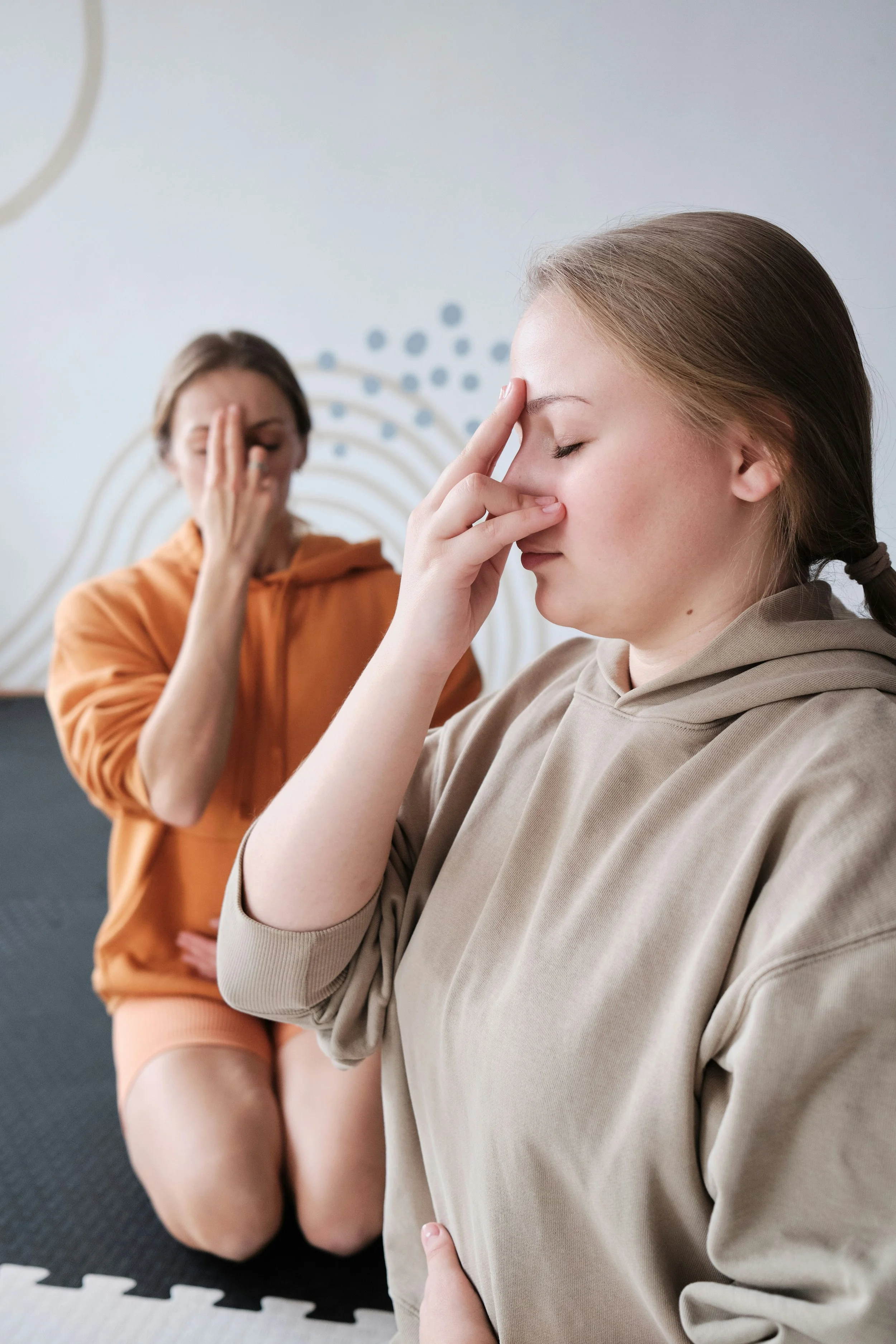 Zwei Frauen in einer Meditationsübung mit geschlossenen Augen, eine sitzt im Vordergrund, die andere im Hintergrund.