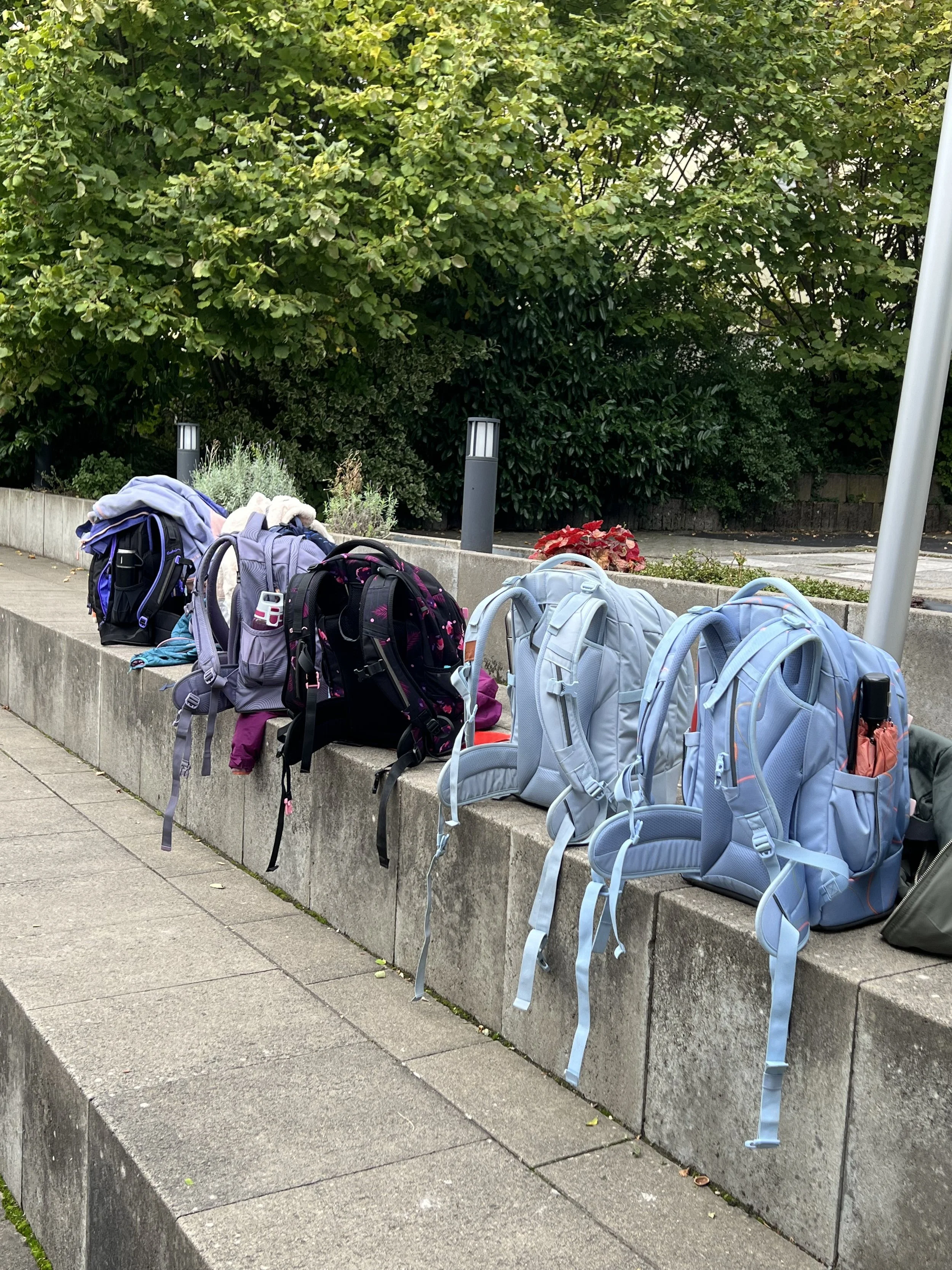 Mehrere Rucksäcke auf einer Steinbank im Freien, im Hintergrund Bäume und eine Beleuchtungssäule.