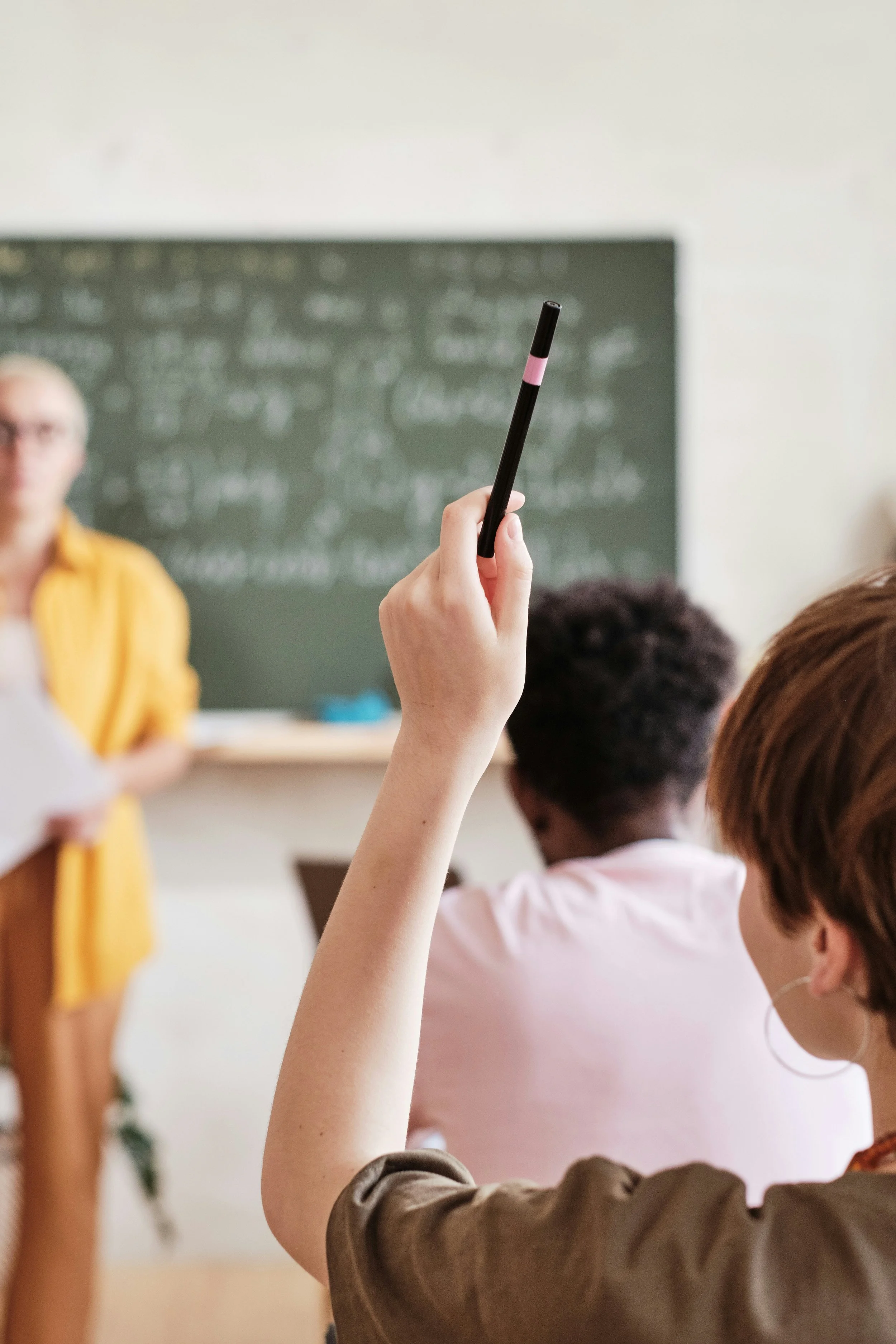Schülerin im Klassenraum hebt die Hand mit Bleistift, während die Lehrerin vorne steht, vor einer Tafel.