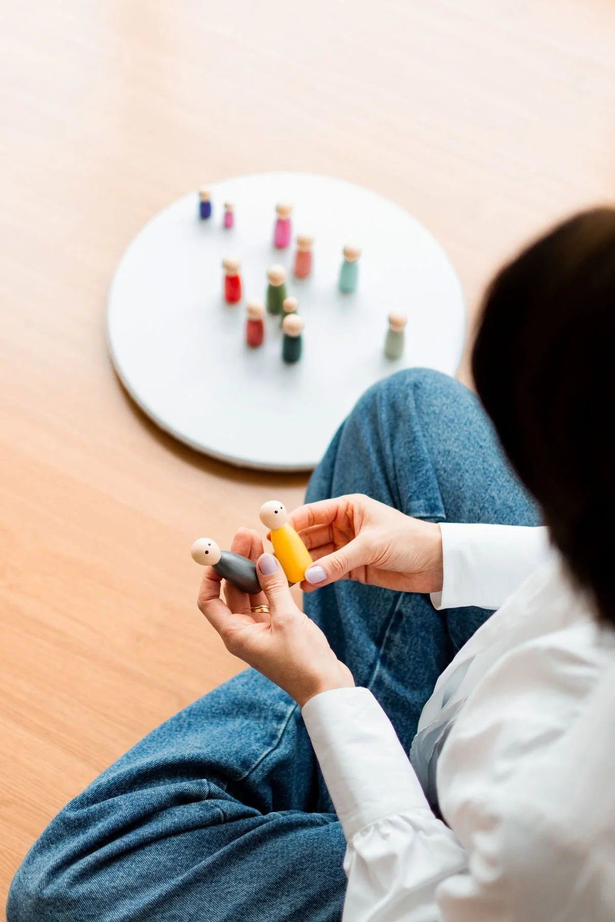 Person, die kleine bunte Holzfiguren hält, während weitere Figuren auf einem weißen Kreis in der Nähe auf dem Boden stehen.