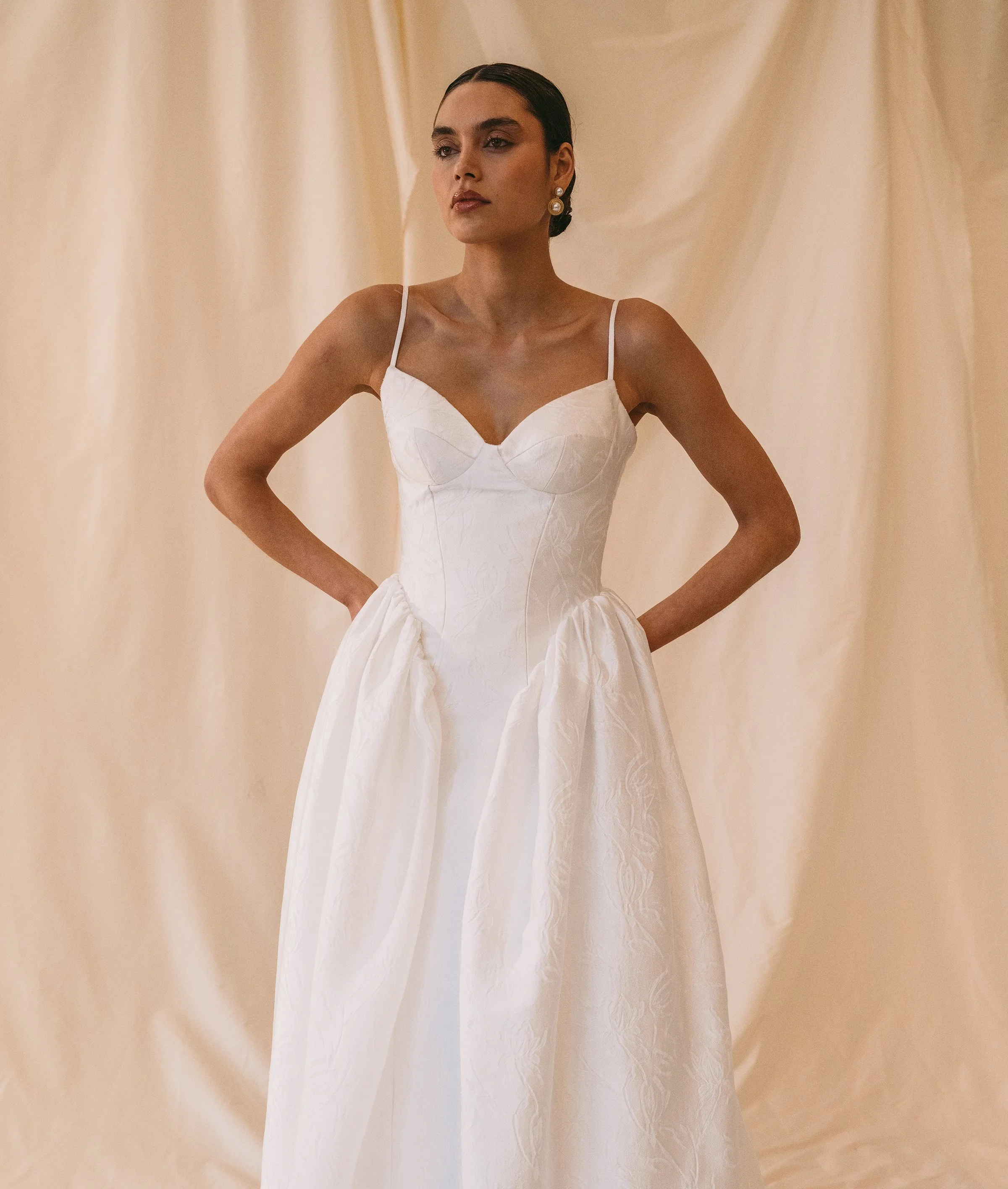 A woman wearing a white bridal gown with spaghetti straps, standing against a cream-colored satin backdrop.