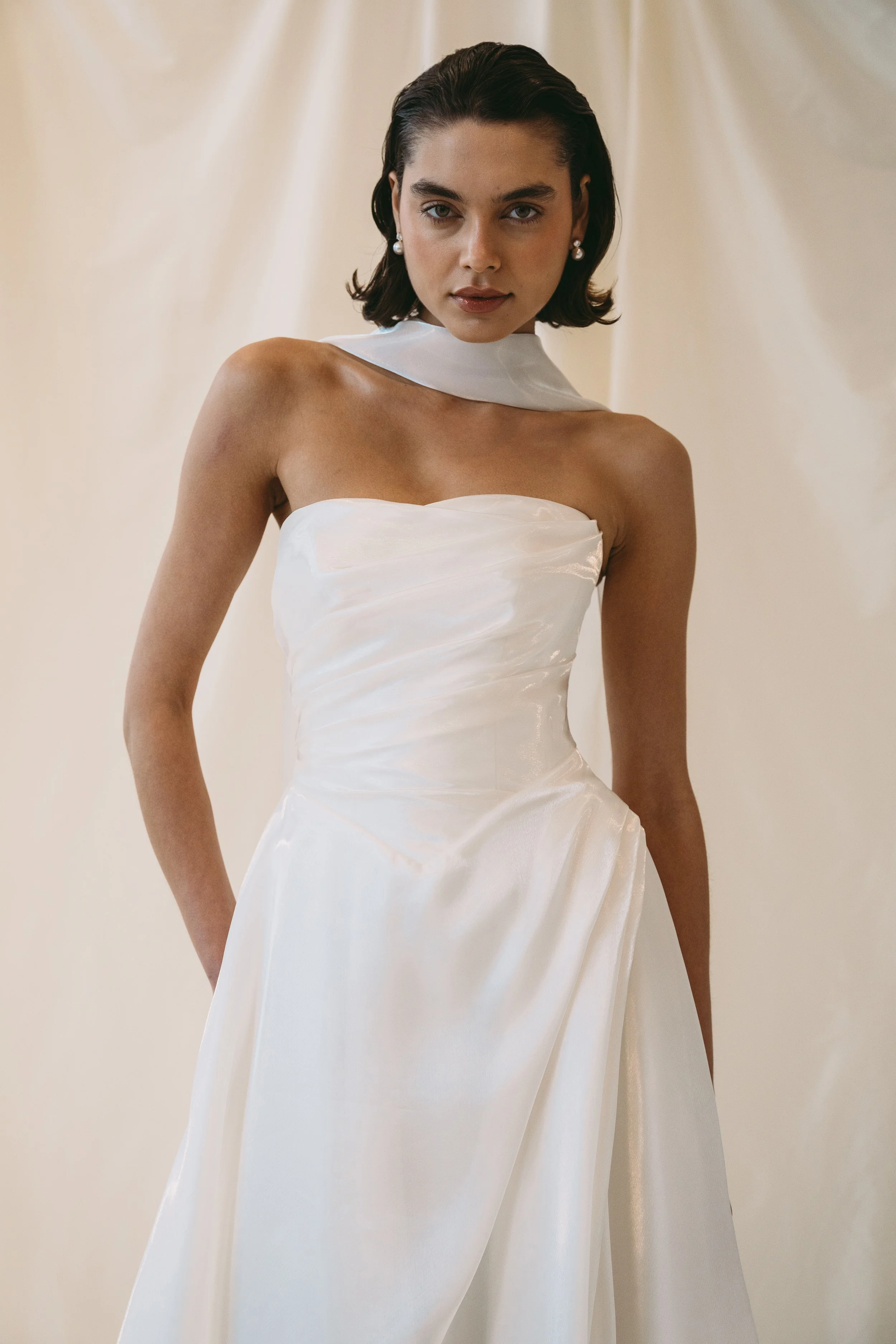Woman wearing a strapless white wedding dress with a high neckline and pearl earrings, standing against a light background.