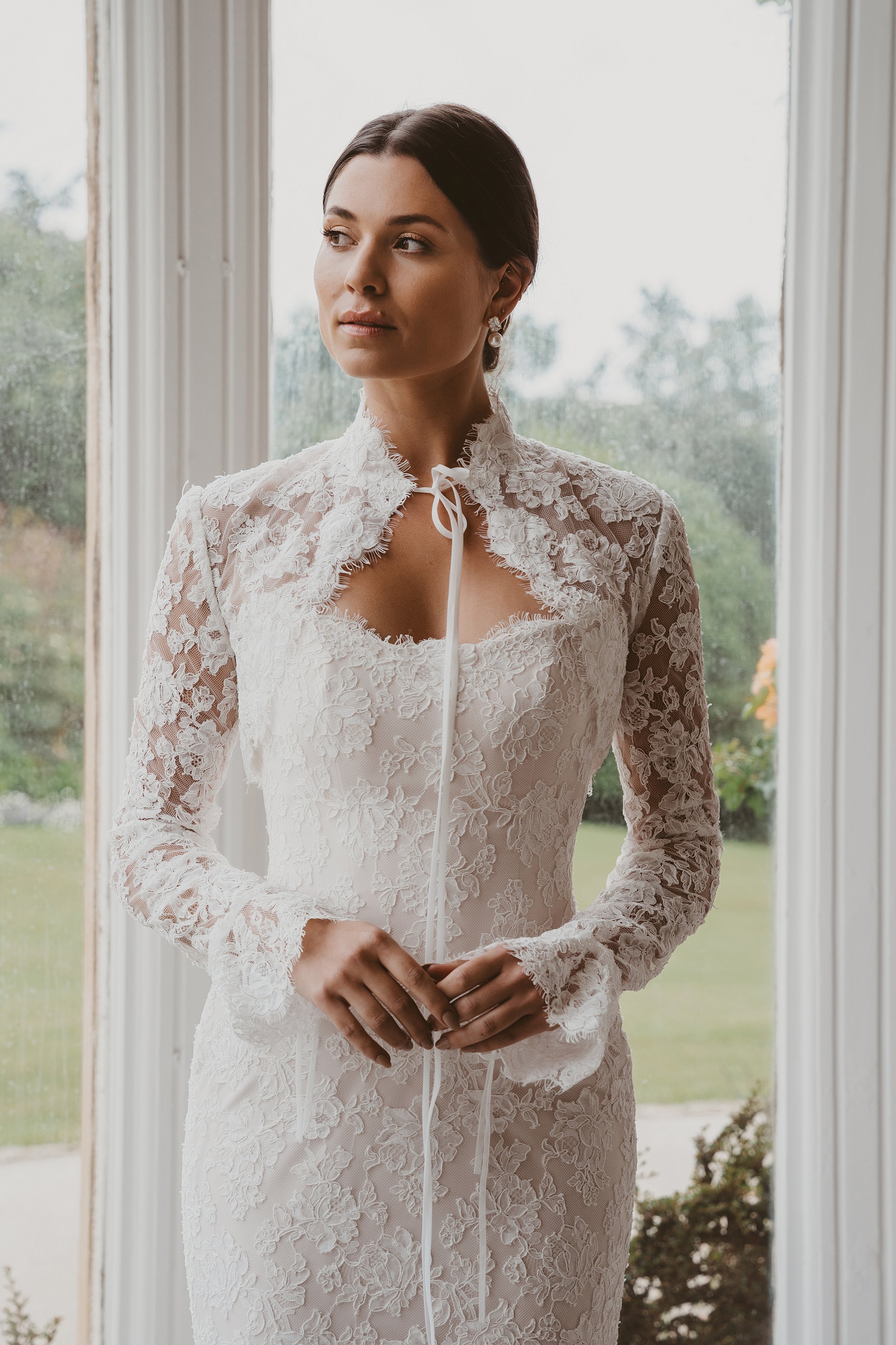 A woman wearing a white bridal gown with spaghetti straps, standing against a cream-colored satin backdrop.