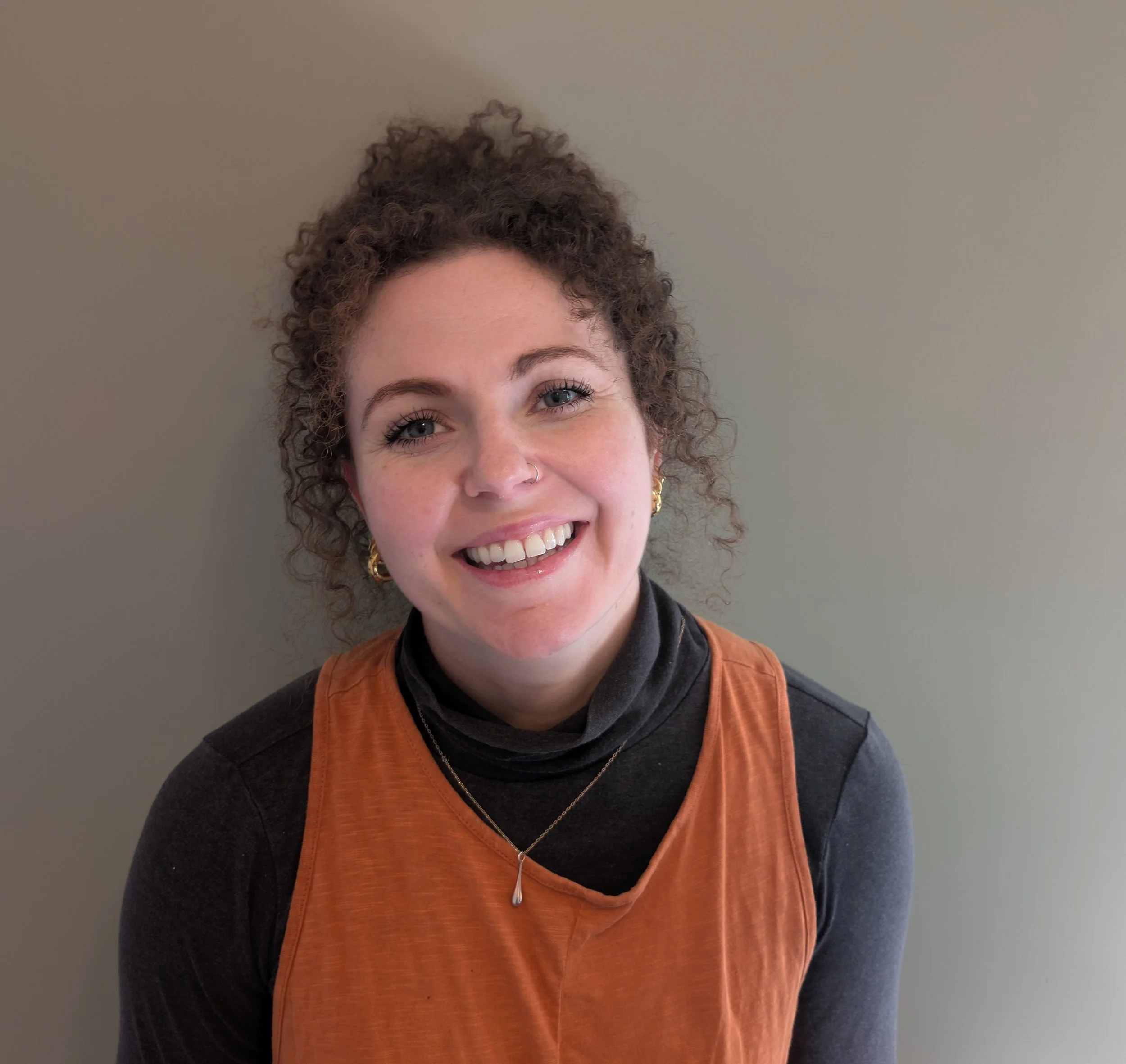 A young woman with curly brown hair smiling, wearing gold earrings, a nose ring, a black turtleneck, an orange sleeveless top, a silver necklace, and standing against a plain wall.