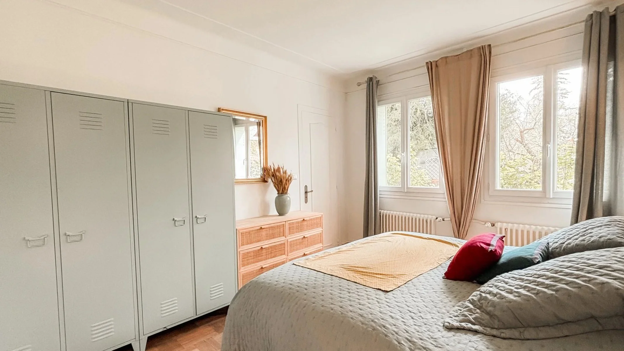 Chambre avec un lit fait, une armoire gris pâle, une commode en bois, un miroir encadré en bois, une plante sèche dans un vase, fenêtres avec rideaux beige et gris, radiateur blanc.