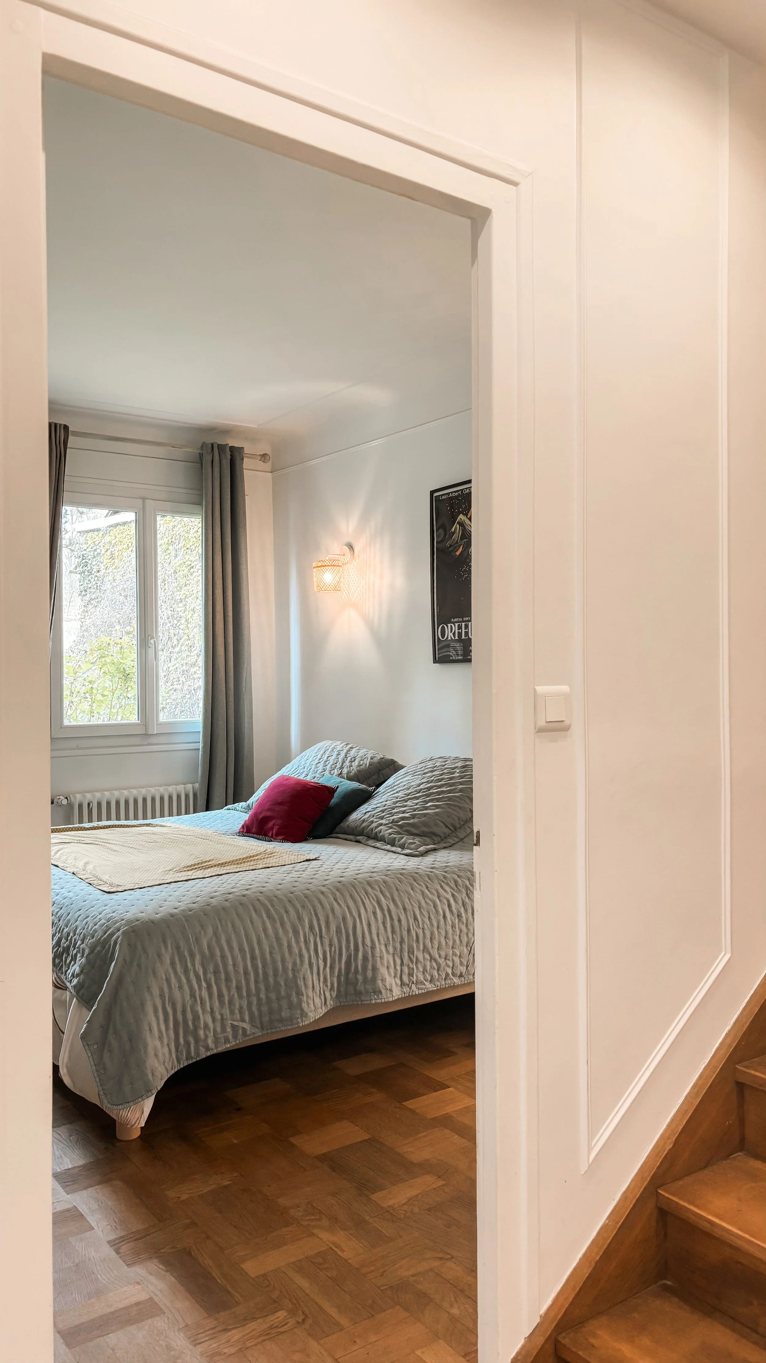 Une chambre vue à travers une porte ouverte, avec un lit, des coussins, une fenêtre avec rideaux, et un mur décoré avec une affiche.