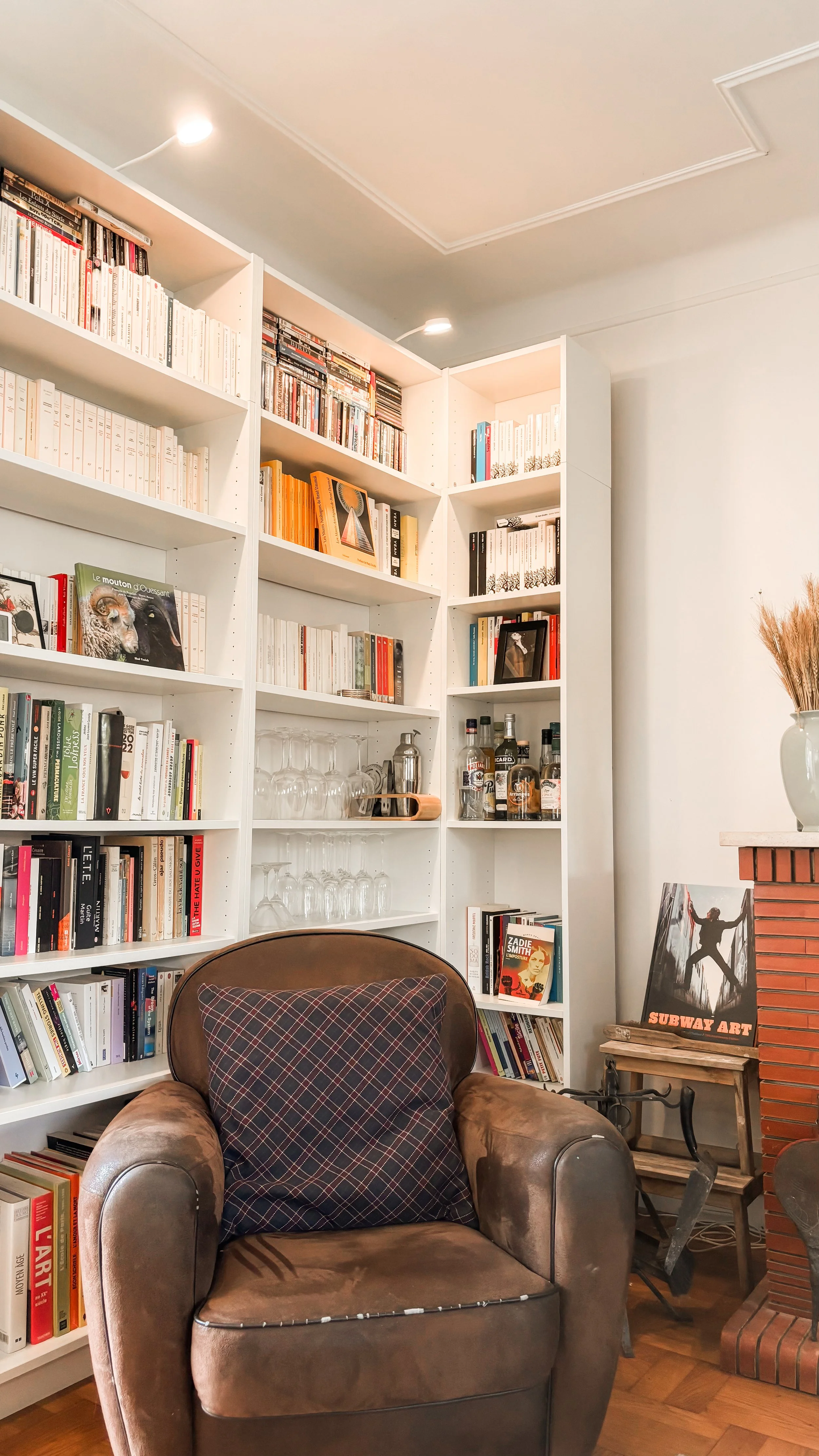 Un coin lecture dans un intérieur moderne avec une bibliothèque blanche remplie de livres, un fauteuil en cuir brun avec un coussin à carreaux, une petite table en bois, et une cheminée en briques. Sur la table, une œuvre d'art intitulée « Subway Art » est visible.
