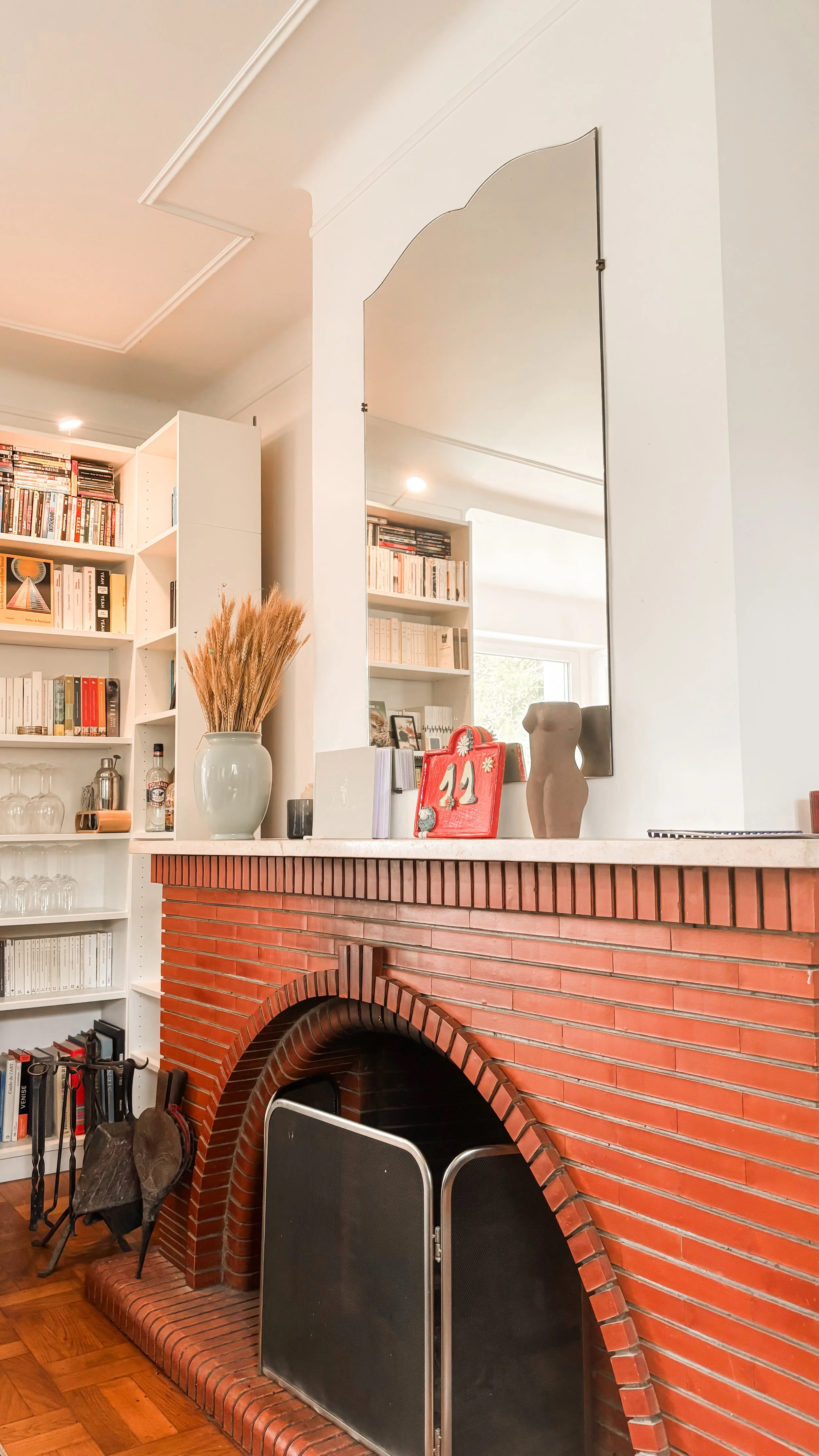 Cheminée en brique rouge avec un pare-feu noir, décorée avec une grande vase blanche contenant de la paille, un calendrier rouge, une sculpture de femme en argile, et un petit livre, dans un intérieur avec étagères pleines de livres.