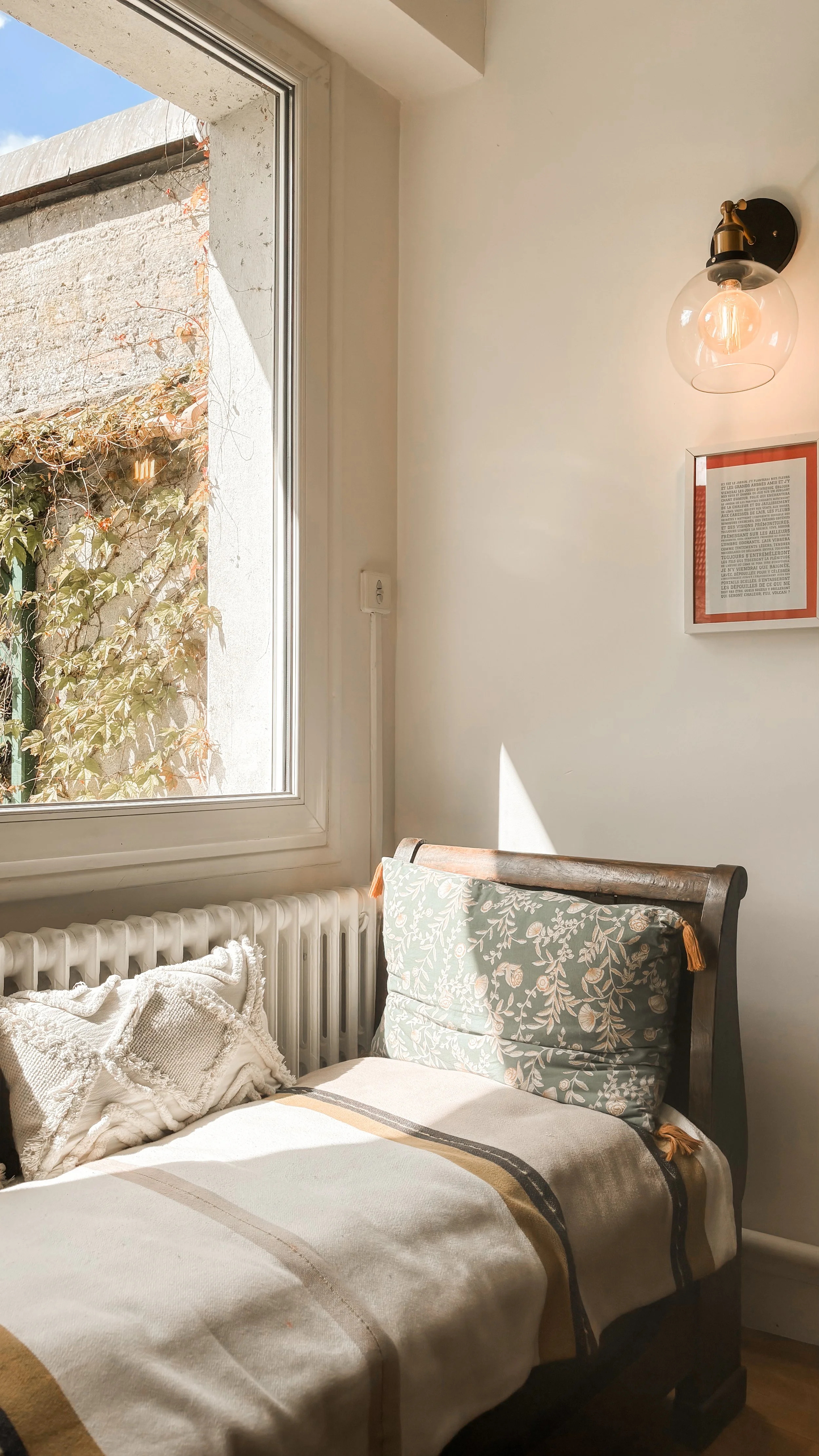 Chambre avec un lit en bois, un coussin floral, une fenêtre laissant entrer la lumière naturelle, un mur blanc avec une lampe et un tableau.