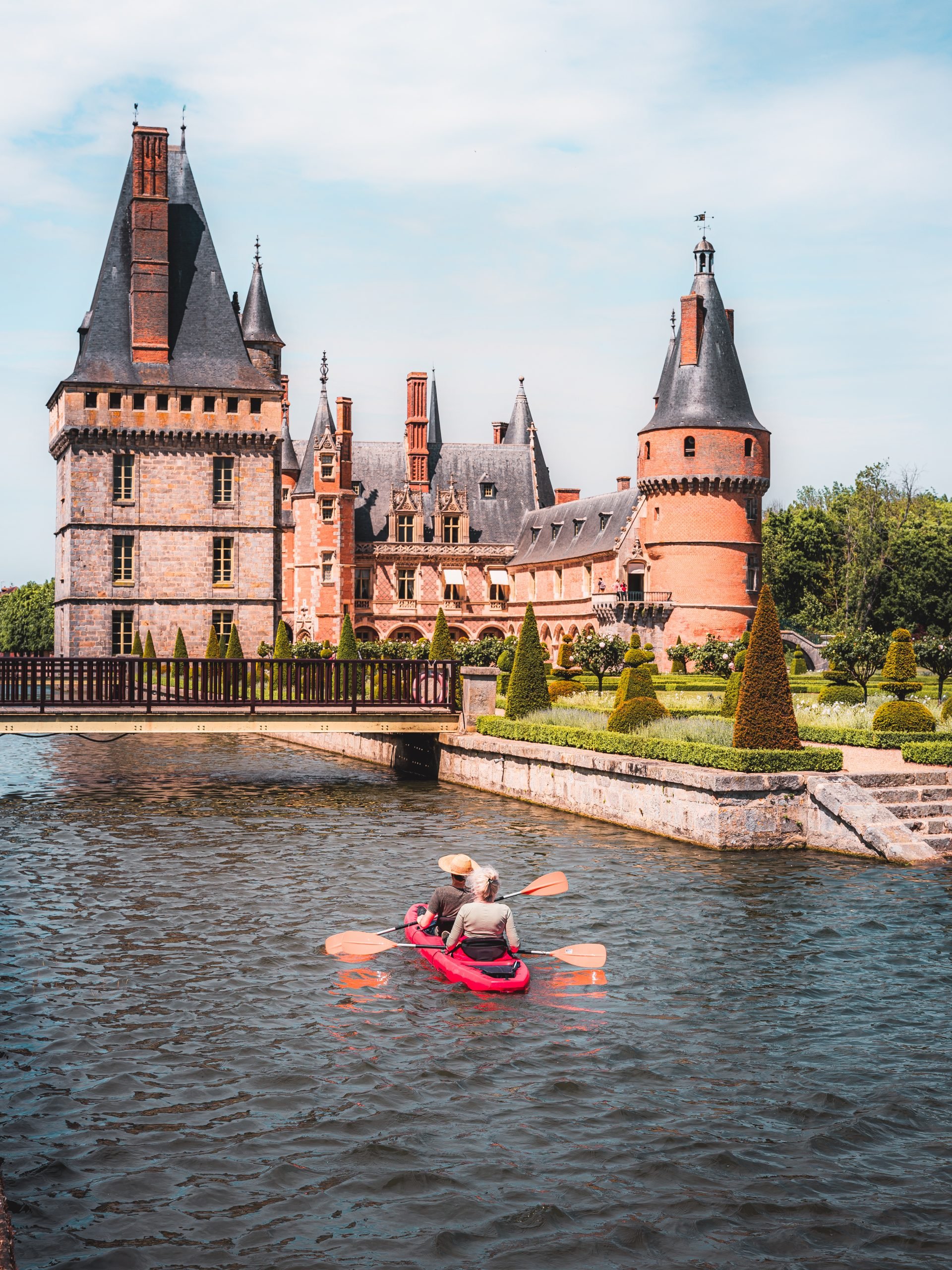 Bateau avec deux personnes paddlant sur un plan d'eau devant un château en pierre avec des tours en ardoise, entouré de jardins bien entretenus.