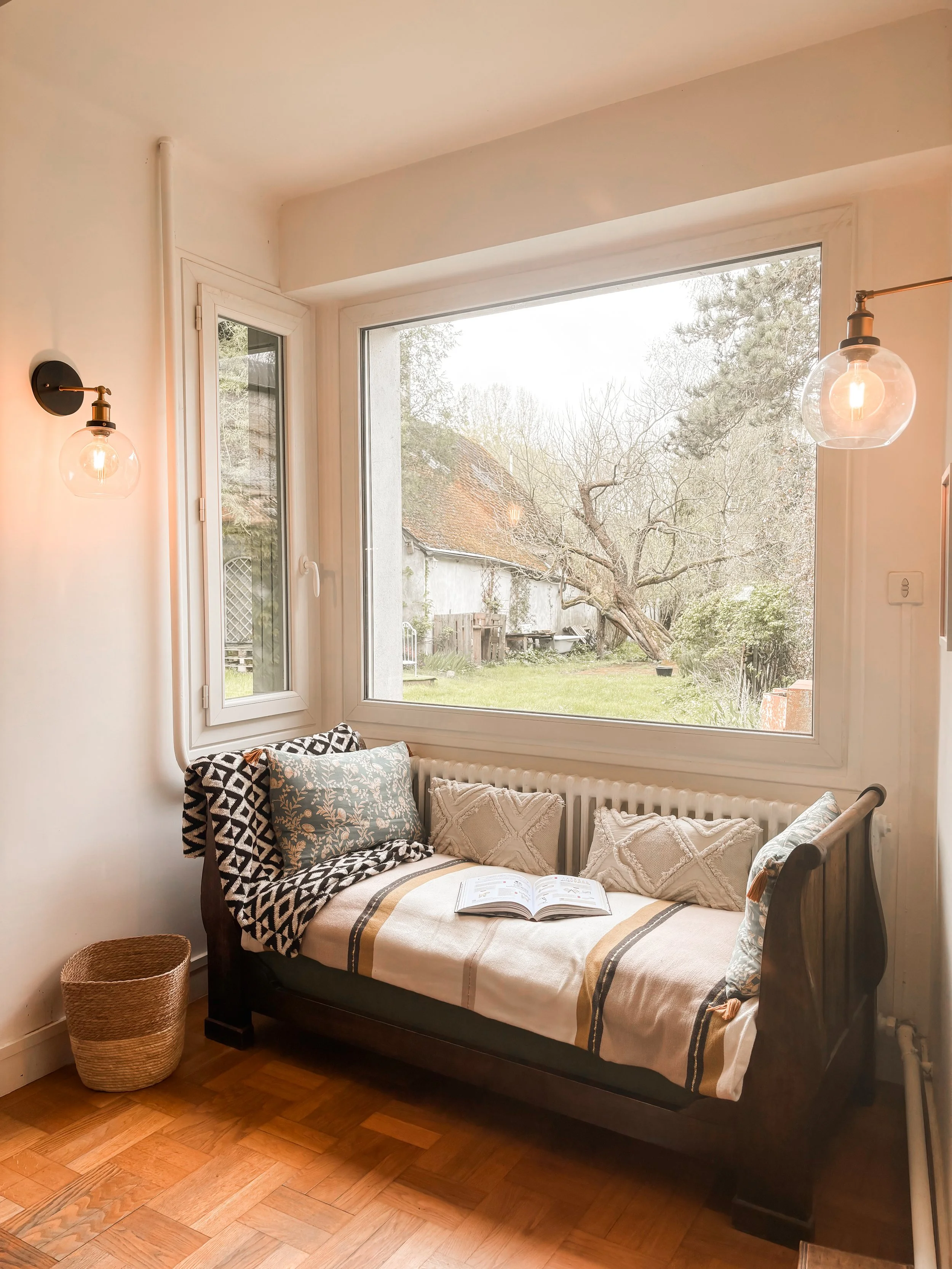 Coin salon avec un canapé-lit en bois noir, plusieurs coussins, une fenêtre large laissant voir un jardin avec un arbre et une clôture, éclairage mural avec deux luminaires en verre.