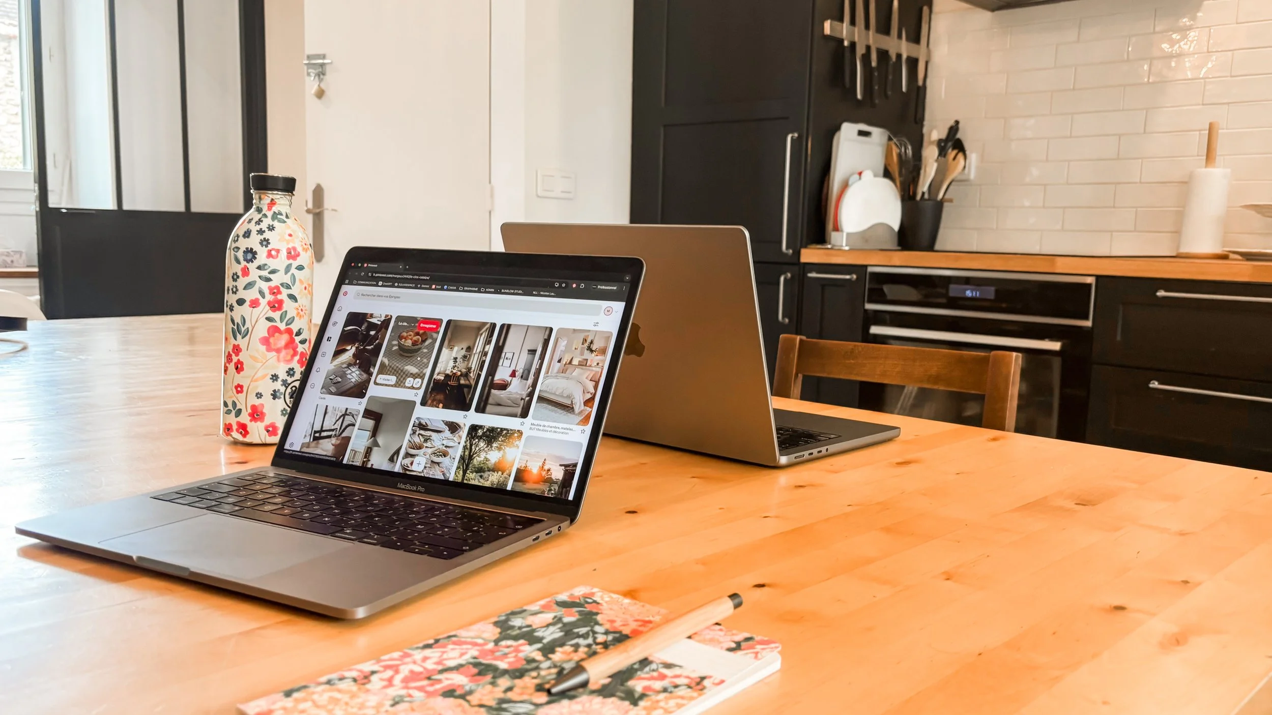 Deux ordinateurs portables ouverts sur une table en bois dans une cuisine moderne, accompagnés d'une bouteille décorative en fleurs, d'un carnet avec un stylo, et un cahier avec un stylo à côté. En arrière-plan, des armoires noires, un plan de travail en bois, et quelques ustensiles de cuisine.