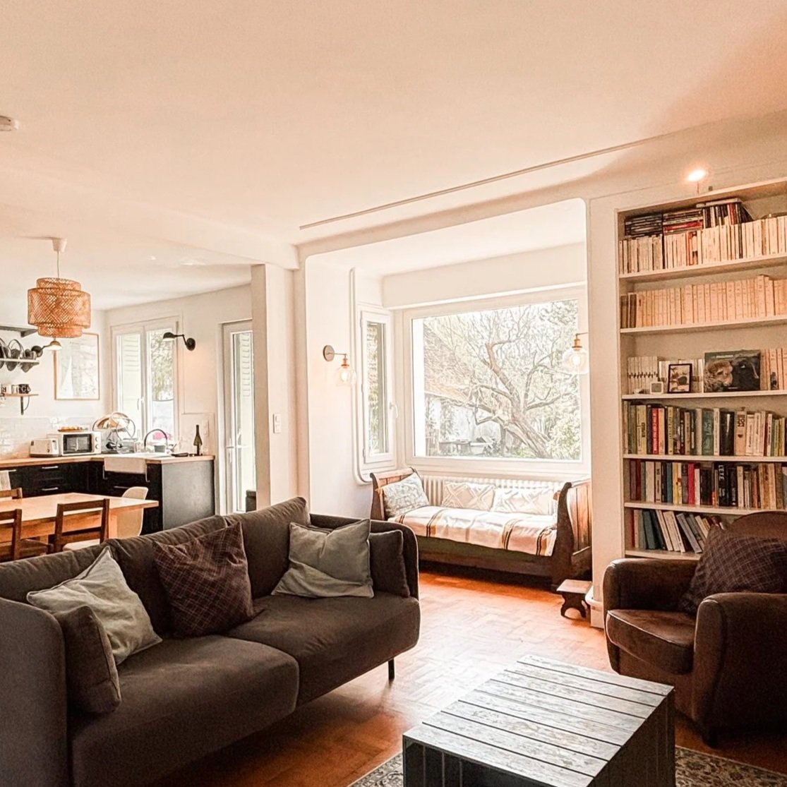 Salon spacieux avec canapé marron, fauteuil, grande bibliothèque, baie vitrée laissant entrer la lumière naturelle, vue sur un arbre