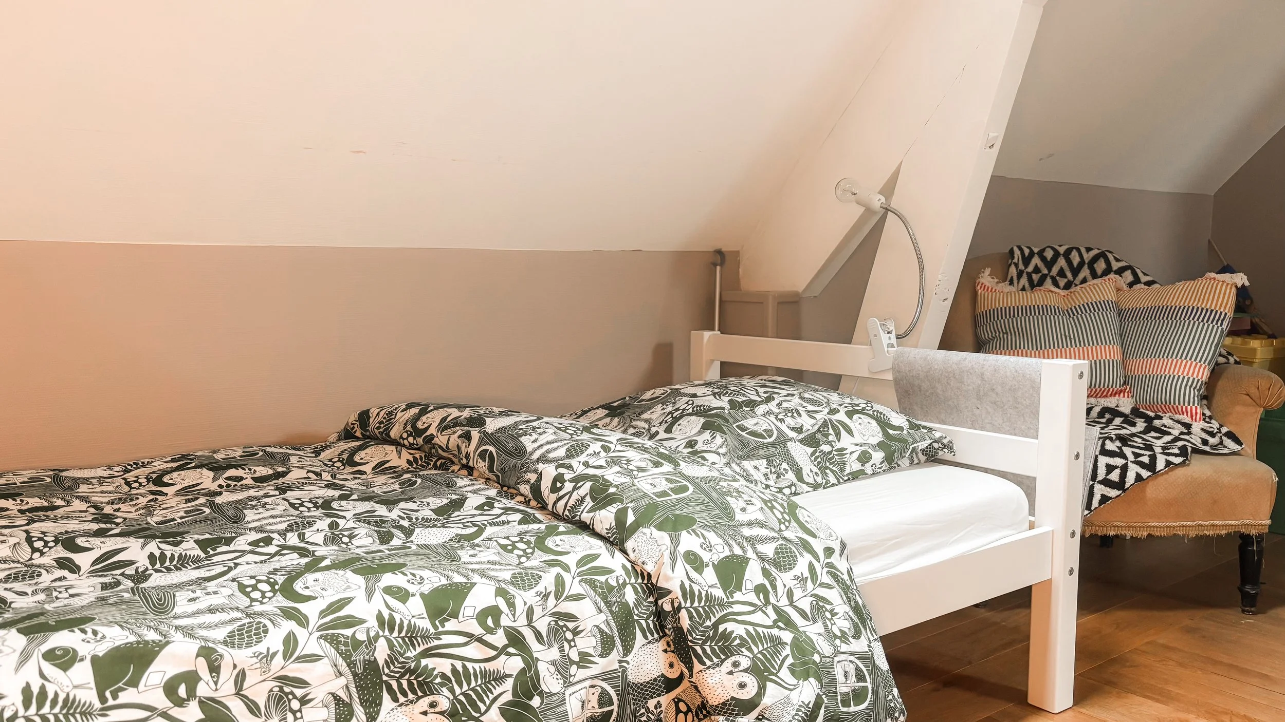 Lit avec une couverture à motifs végétaux, près d'un mur blanc sous un plafond en pente, avec une tête de lit beige et plusieurs coussins colorés et motifs variés.