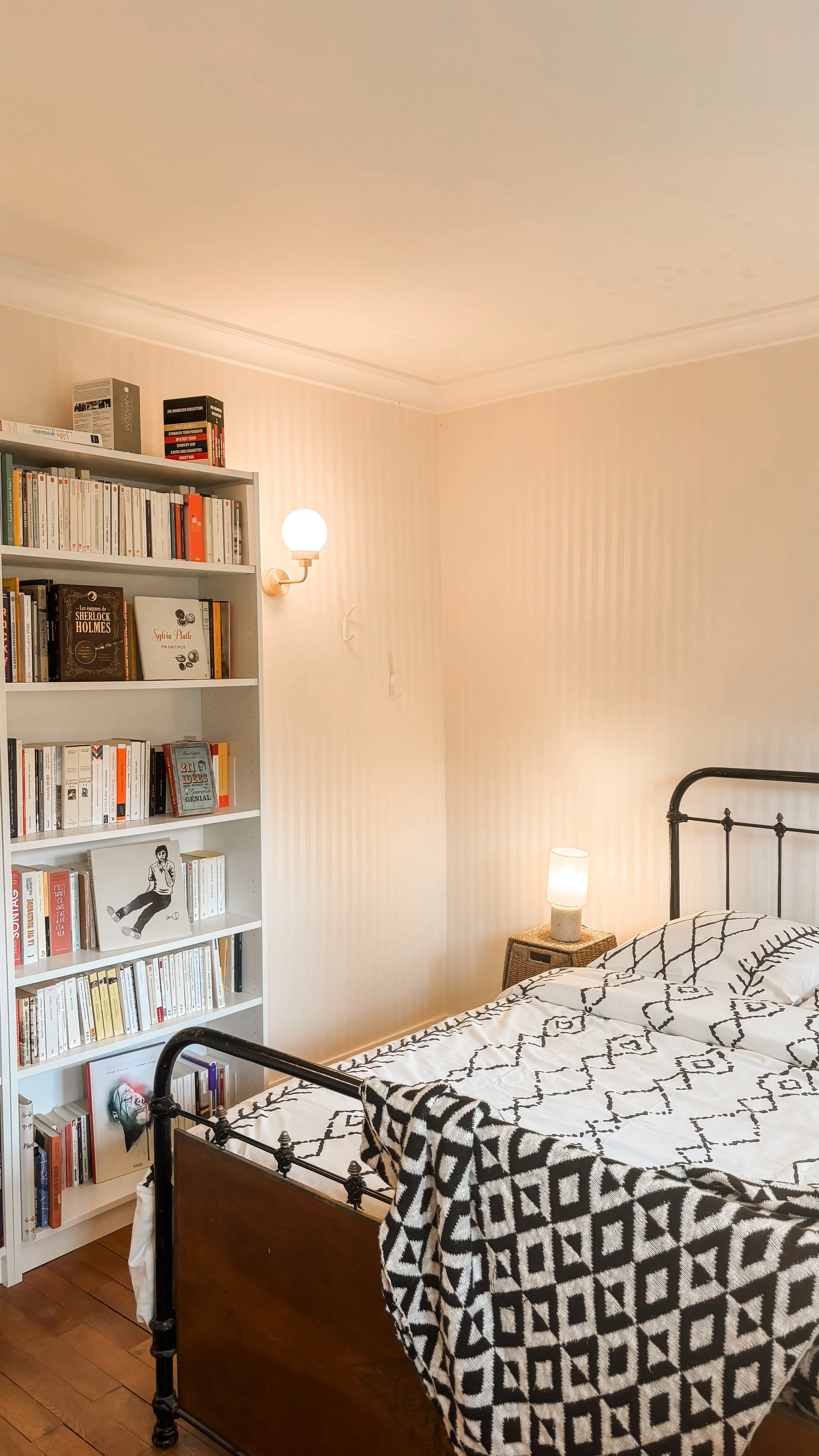 Chambre avec un lit en fer noir, une table de chevet en rotin avec une lampe, une étagère blanche remplie de livres, et un mur à rayures jaunes pâles.