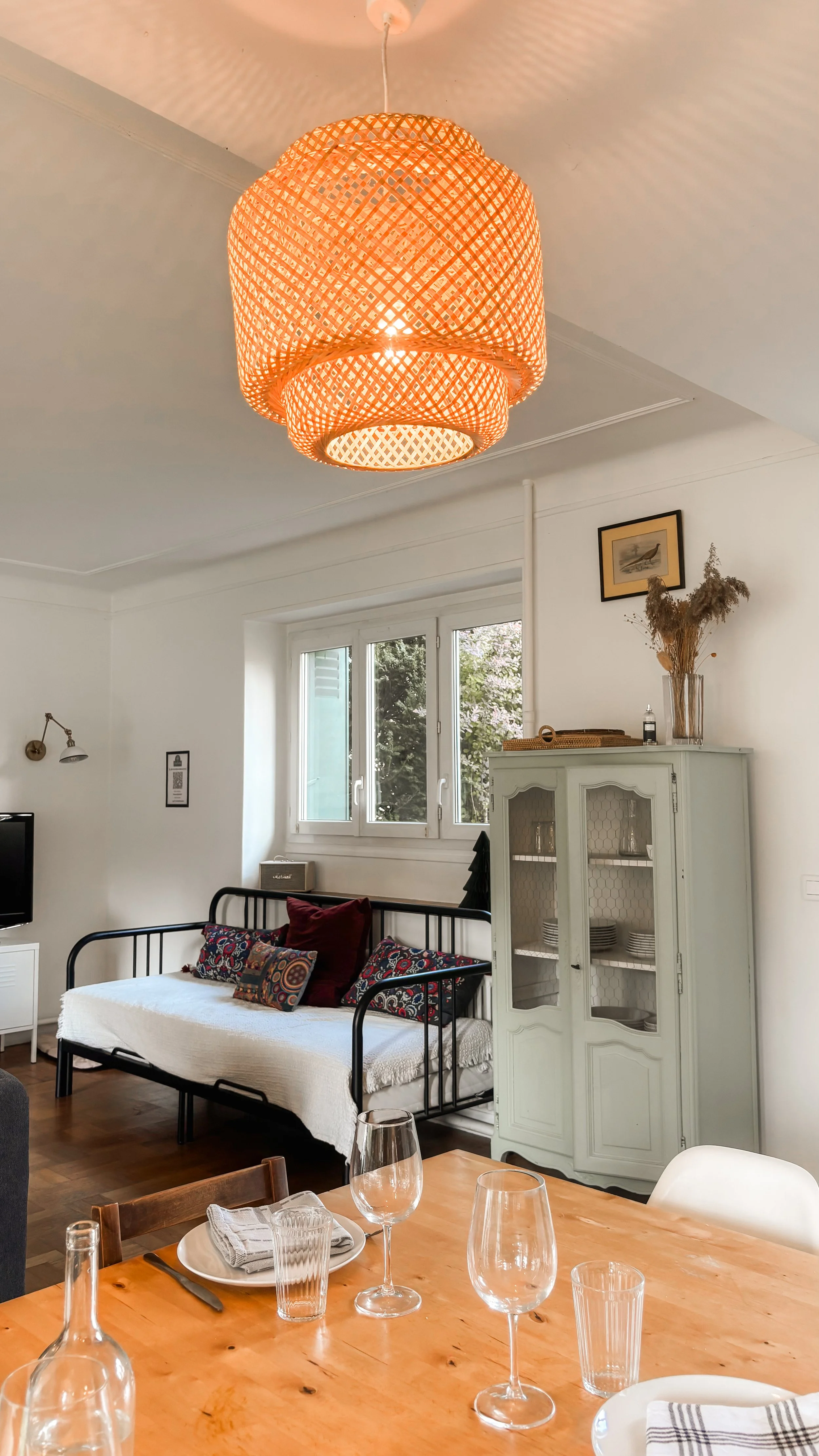 Intérieur d'un salon avec un lustre en rotin suspendu au plafond, une fenêtre laissant entrer la lumière, un canapé noir avec des coussins colorés, une armoire en bois peint en blanc avec des décorations et des plantes séchées sur le dessus, et une table en bois avec des verres à vin et des assiettes.