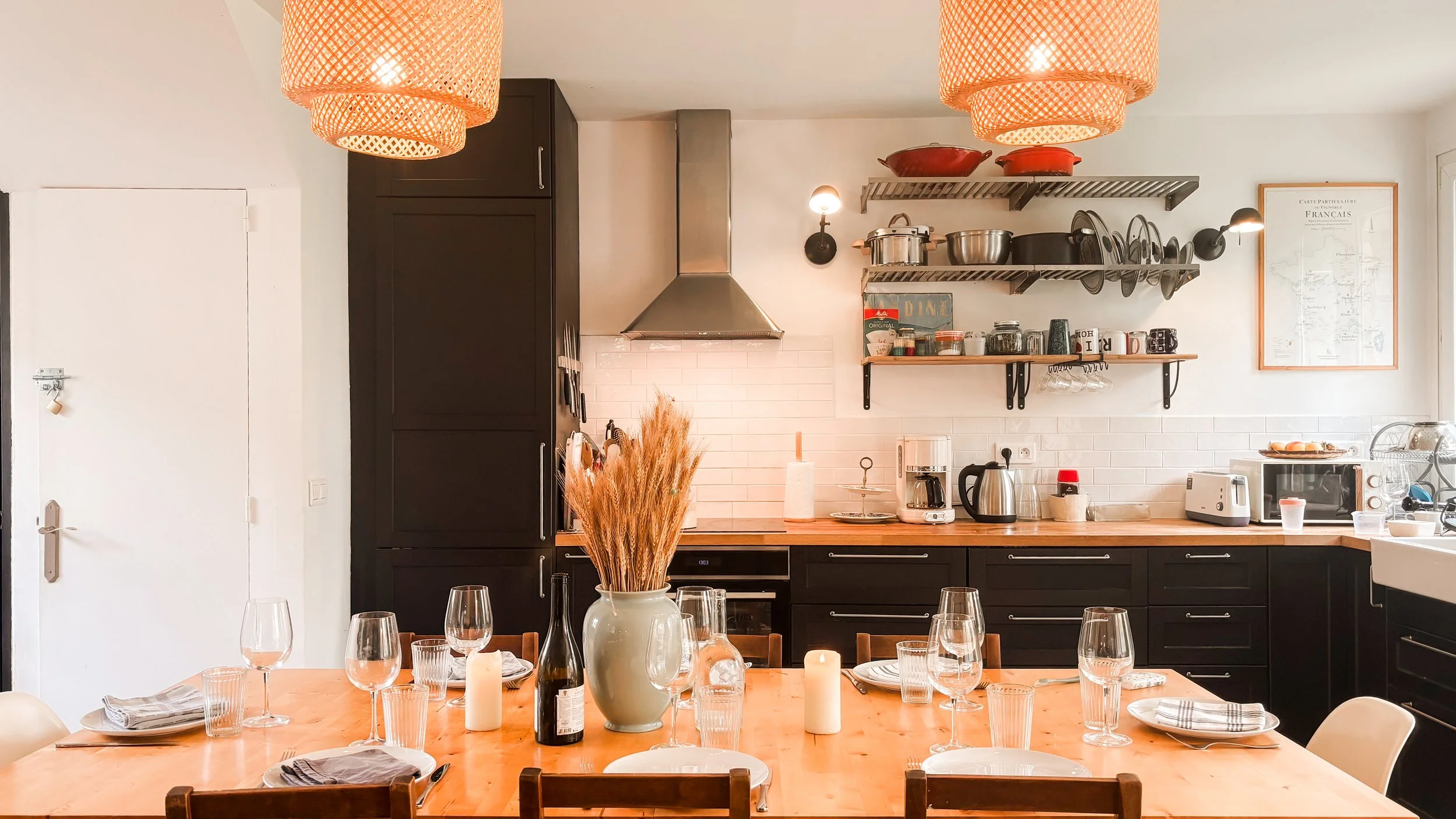 Cuisine moderne avec étagères en métal, casseroles rouges, micro-ondes, cafetière, verres et assiettes sur une table en bois, éclairage chaleureux, décor minimaliste.