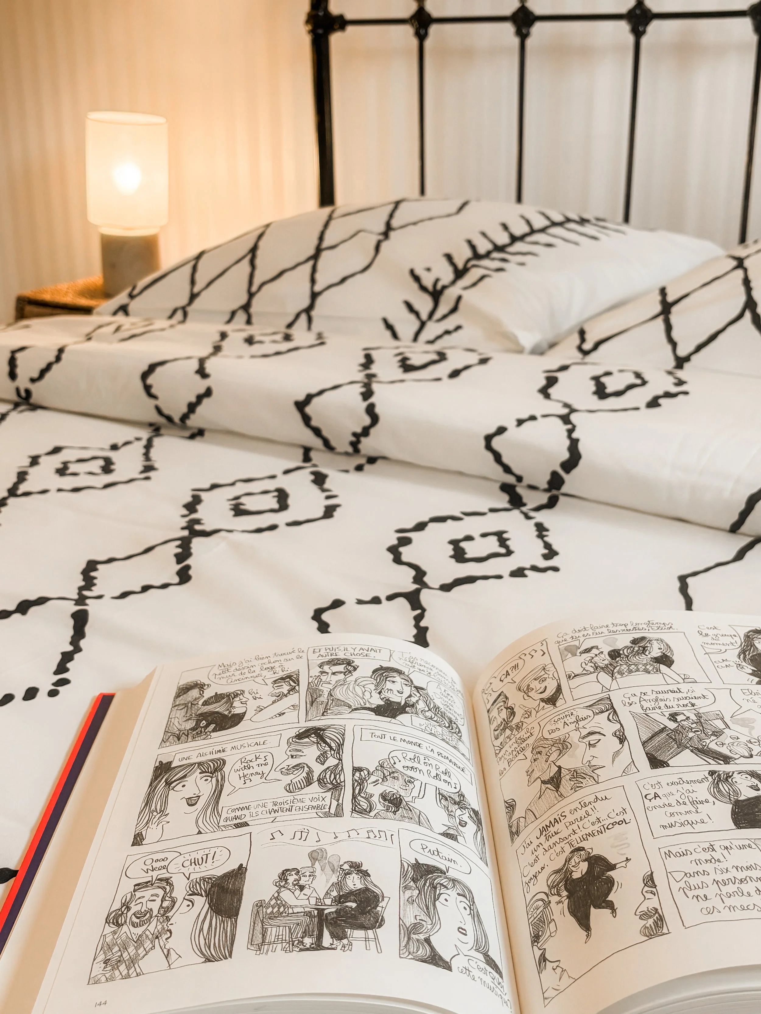 Une chambre avec un lit à cadre en métal noir, des coussins à housses blanches avec un motif noir, une table de chevet avec une lampe et un livre ouvert de bandes dessinées en français. La chambre a des murs en papier peint beige et un éclairage chaleureux.