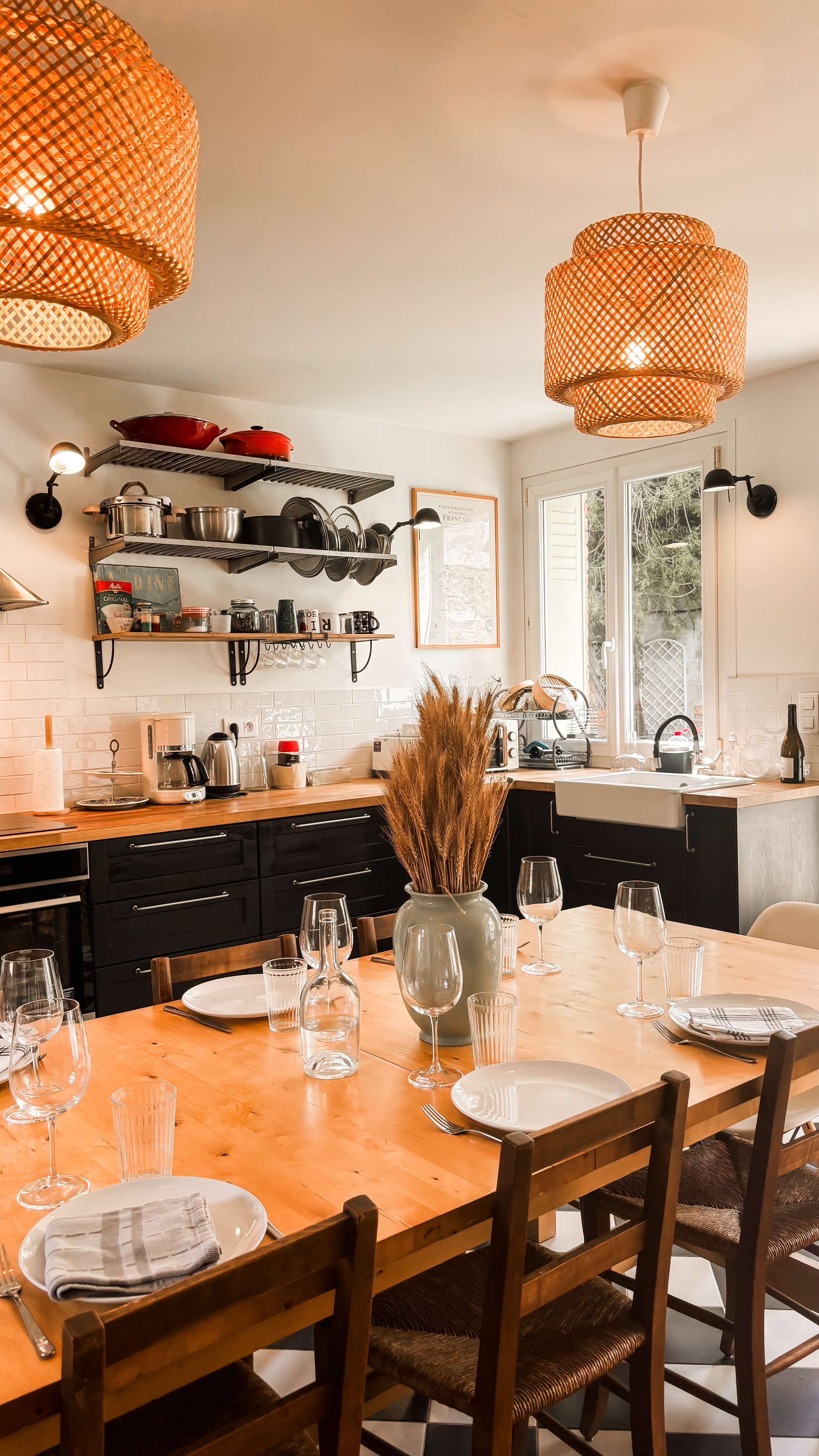 Une salle à manger de style moderne avec un grand table en bois, des verres à vin, des assiettes, des couverts, une nappe en tissu, une grande vase avec des herbes ou des céréales séchées, des chaises en bois, un mur blanc avec des étagères en métal contenant des casseroles, des pots, et des mugs, une fenetre laissant entrer de la lumière naturelle, un éclairage chaud avec des suspensions en rotin