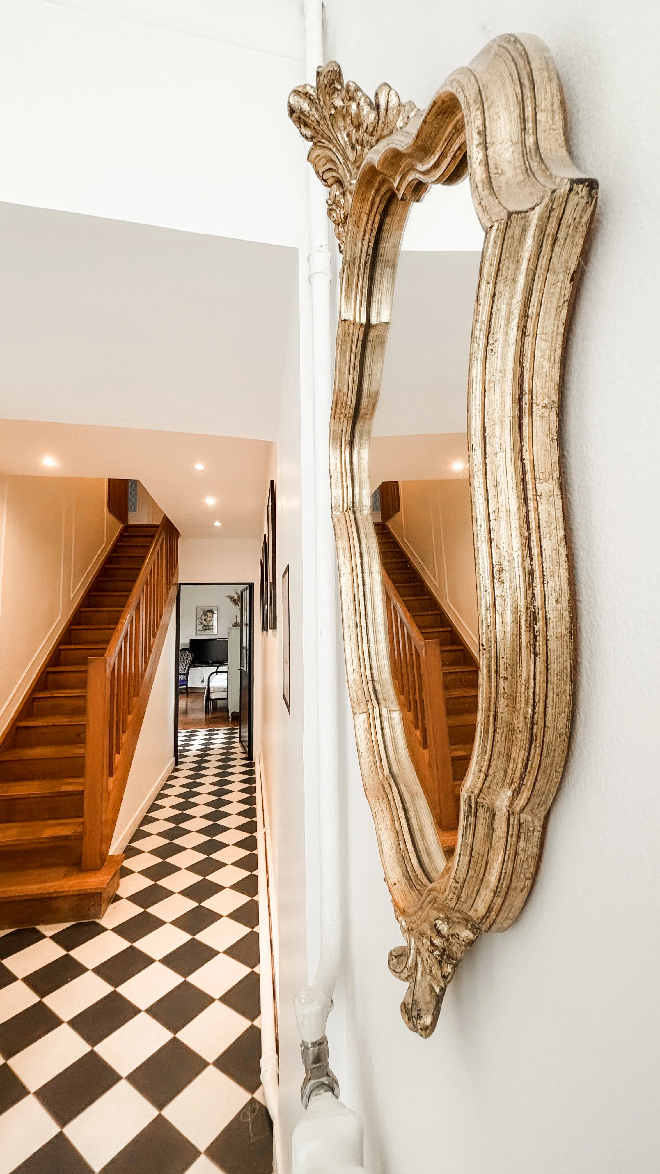 Un miroir antique avec cadre en bois travaillé accroché sur un mur blanc dans un couloir, avec des escaliers en bois à gauche et un sol en carreaux noirs et blancs.