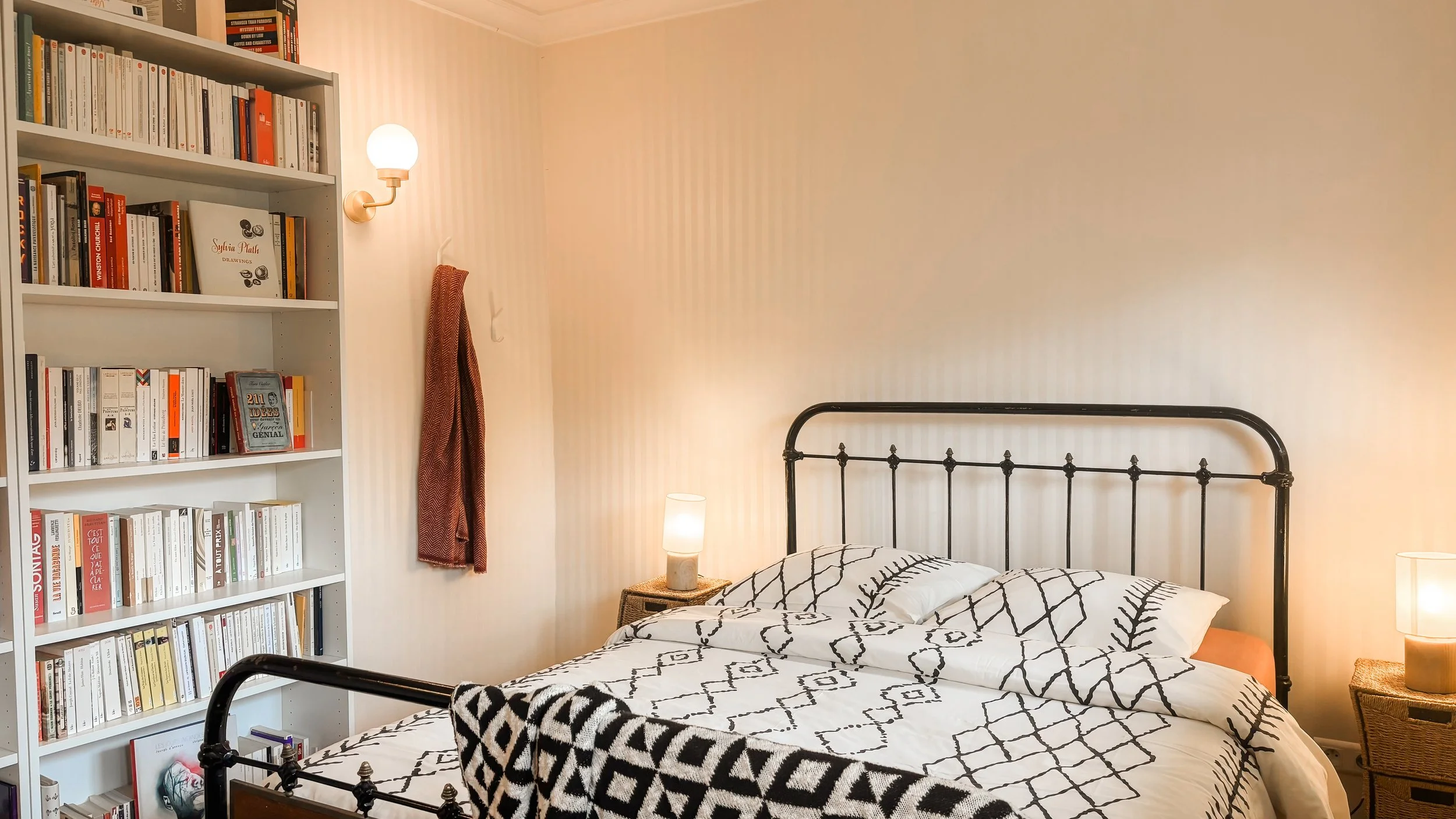 Une chambre avec un lit à tête en fer noir, une bibliothèque remplie de livres blancs, une lampe de chevet sur une table en osier, un mur avec un ruban rouge et une lampe murale, une parure de lit blanche avec des motifs noirs, un autre lampe à côté du lit.