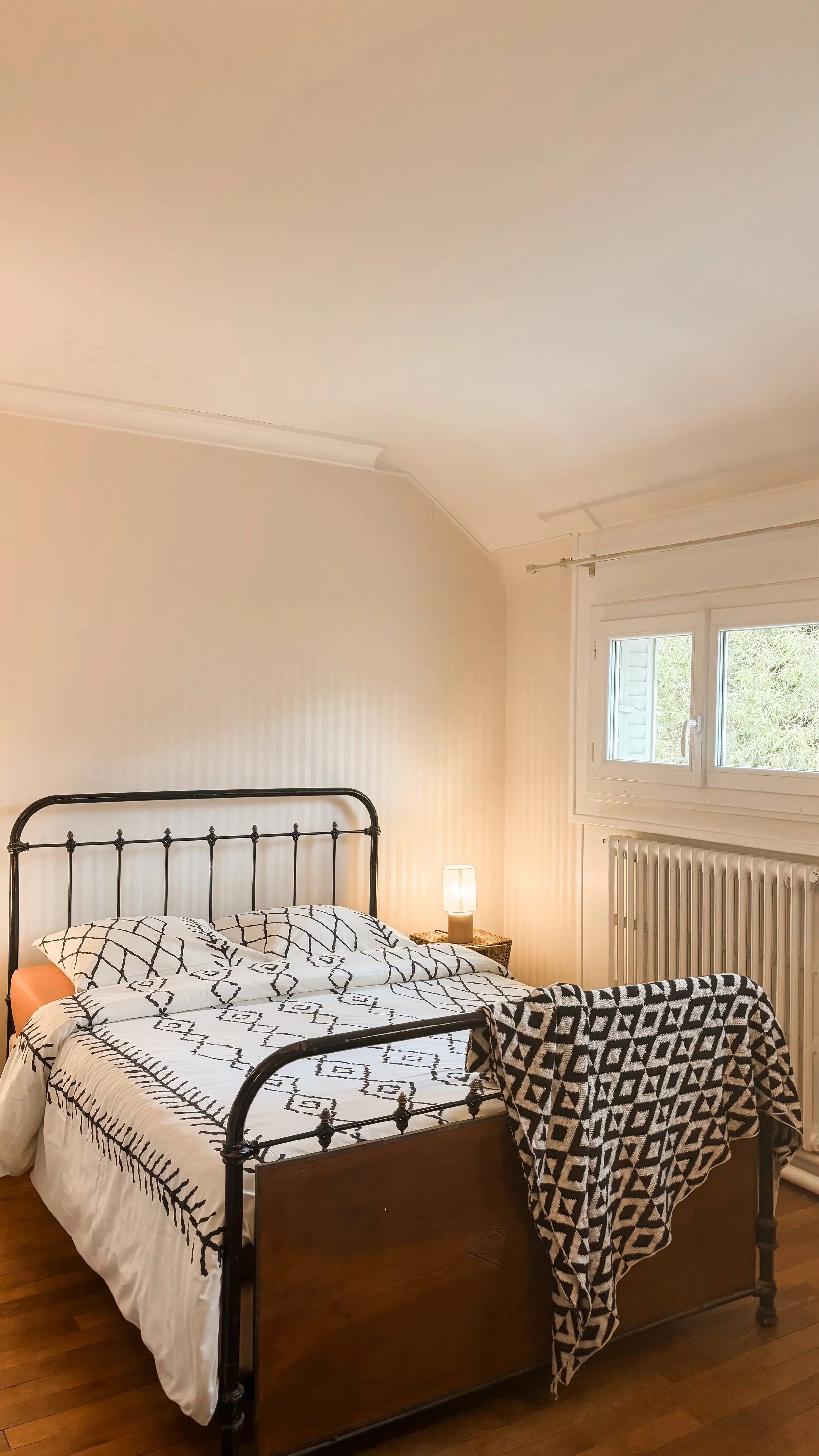 Une chambre avec un lit à cadre en métal noir, drap et coussins à motifs noirs, blancs et beiges, une table de chevet avec une lampe allumée, une fenêtre, un radiateur, et un rideau