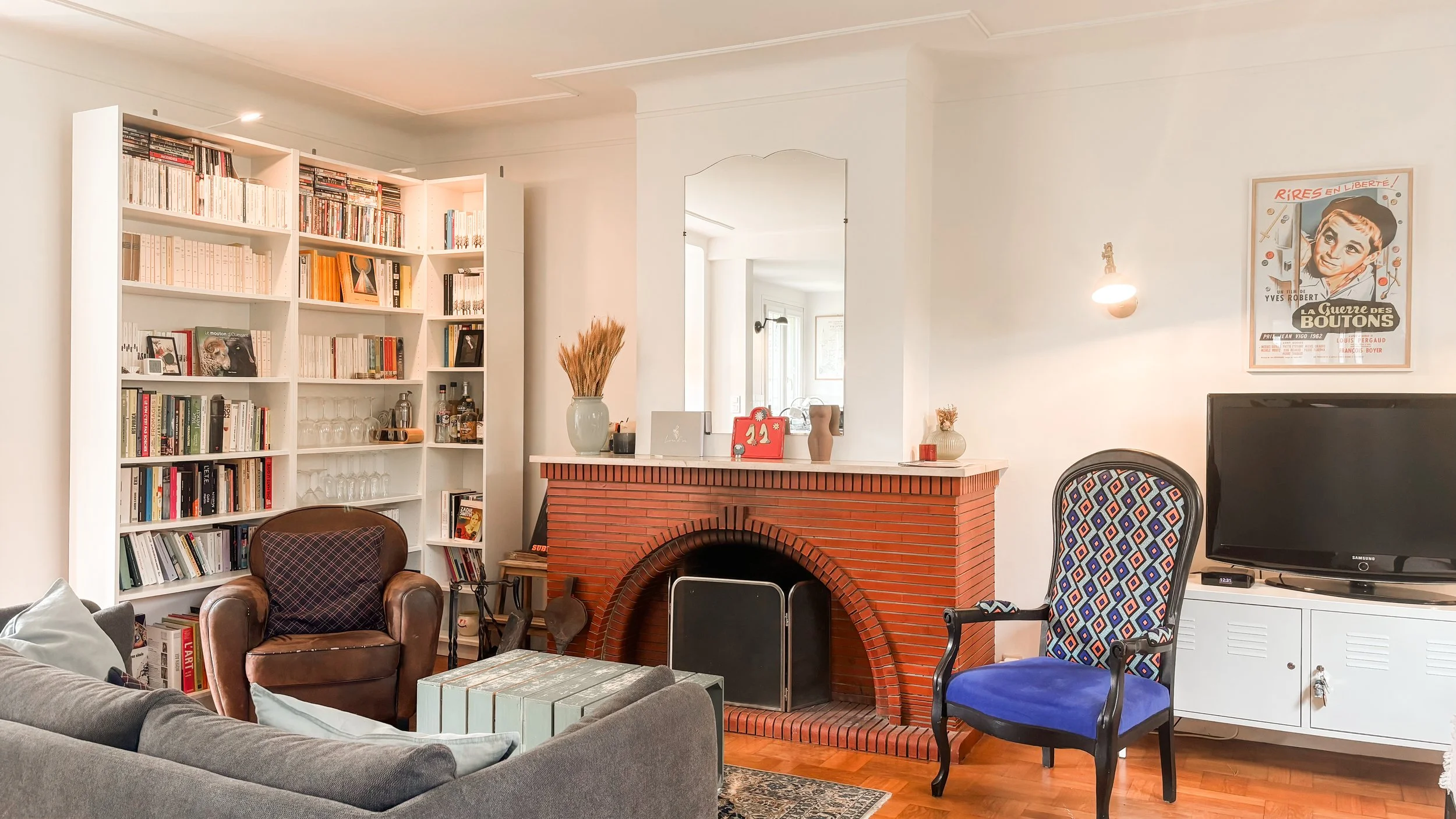 Salon avec cheminée en briques rouges, étagère pleine de livres, fauteuil en cuir marron, canapé gris, table en bois, télévision, peinture vintage sur le mur, décorations variées.