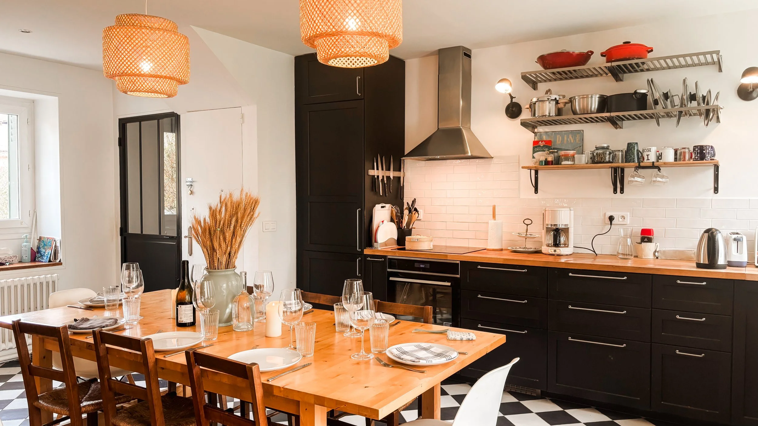 Cuisine moderne avec un mur de carreaux blancs, des étagères en métal avec des casseroles et des mugs, un plan de travail en bois, un placard noir, une hotte en métal, et une grande table en bois prête pour un repas, avec des verres et des assiettes.