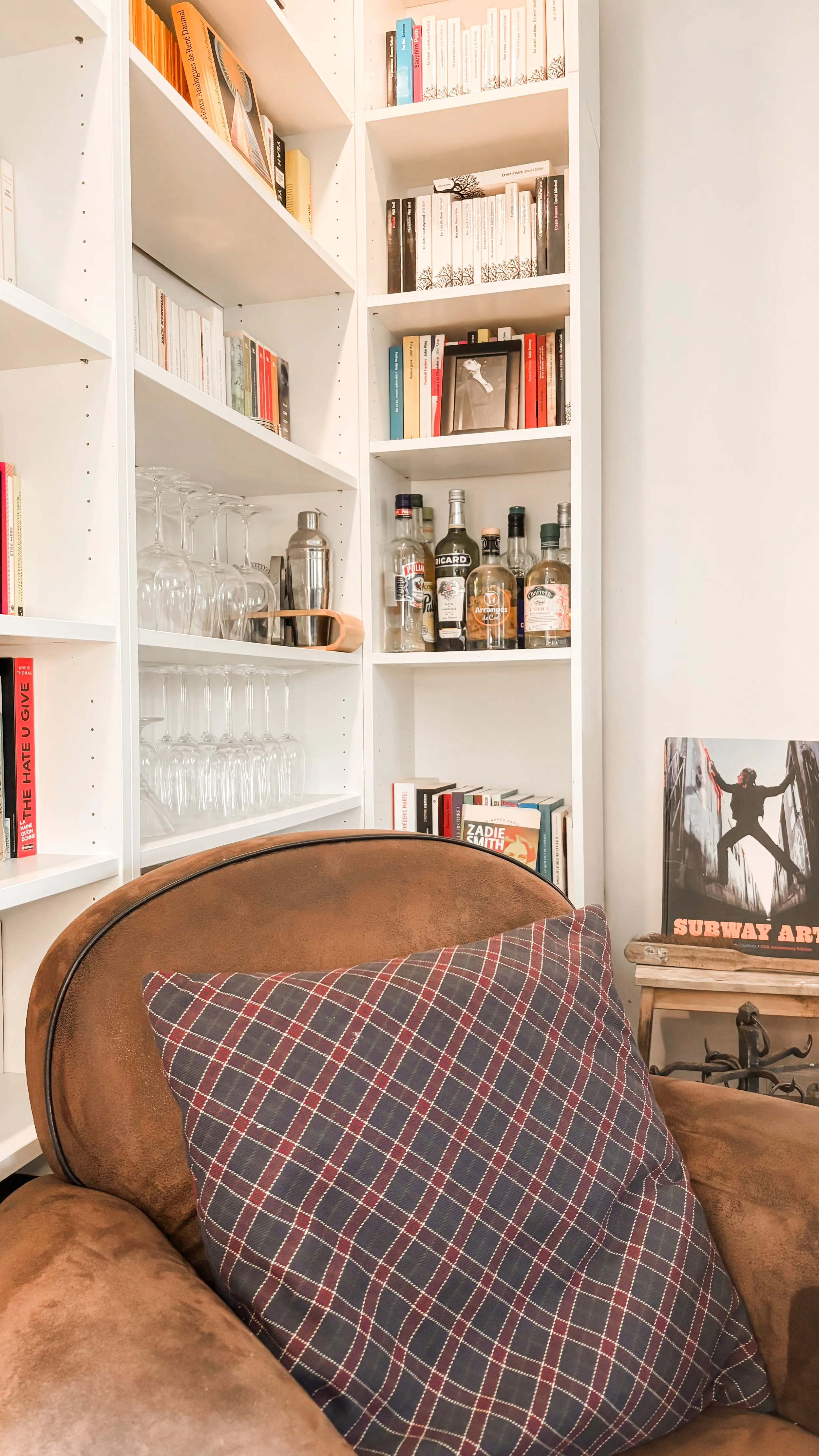 Une étagère blanche avec des livres, des verres à vin, et une bouteille d'alcool. En face, un fauteuil en cuir marron avec un coussin à carreaux. À côté, une affiche de Subway Art avec une silhouette en graffiti.