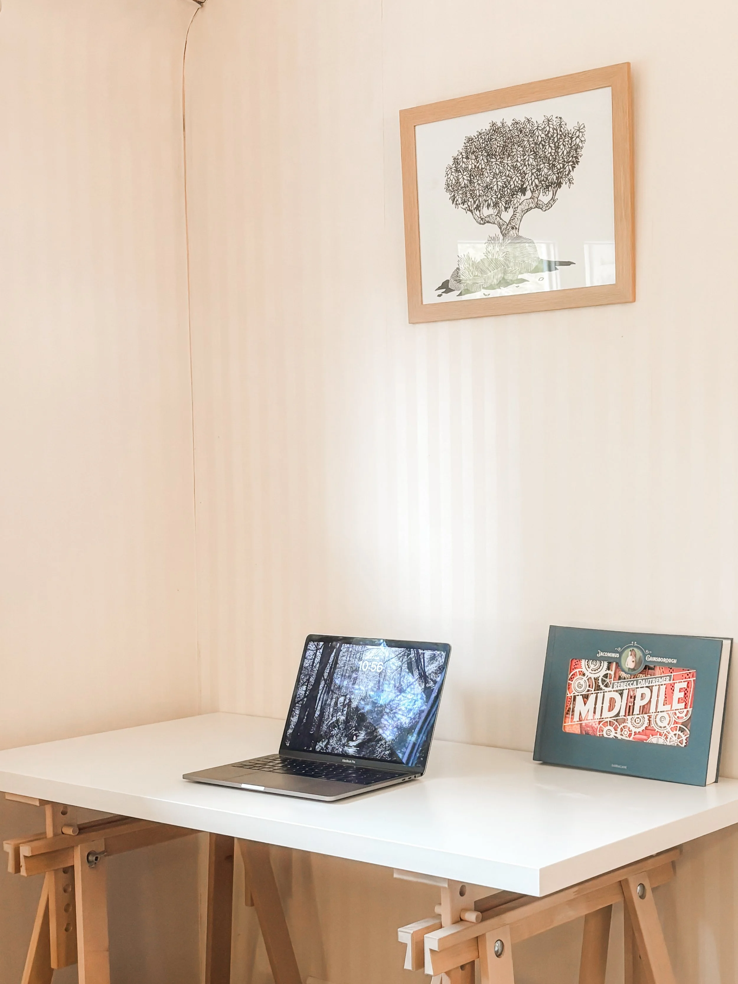 Un ordinateur portable Apple MacBook posé sur un bureau blanc avec une tablette à côté affichant une couverture de livre intitulée 'Middle'. Dans le fond, un mur beige clair et une affiche au mur avec un arbre dessiné.