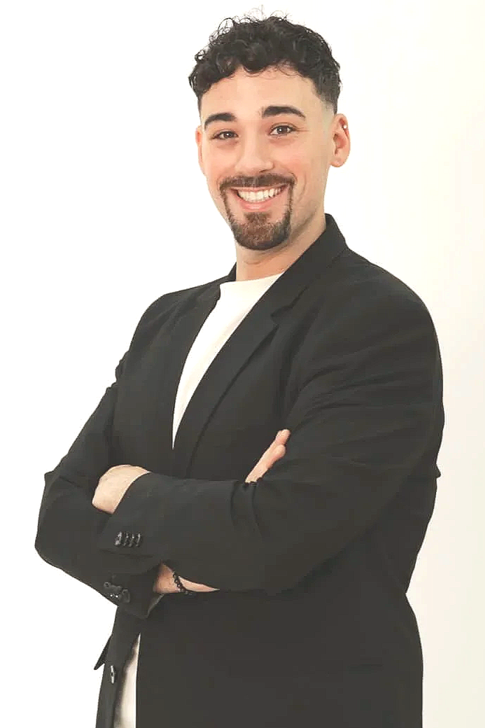 A young man with dark curly hair, a beard, and mustache, smiling and standing with arms crossed against a white background. He is wearing a black blazer over a white shirt.