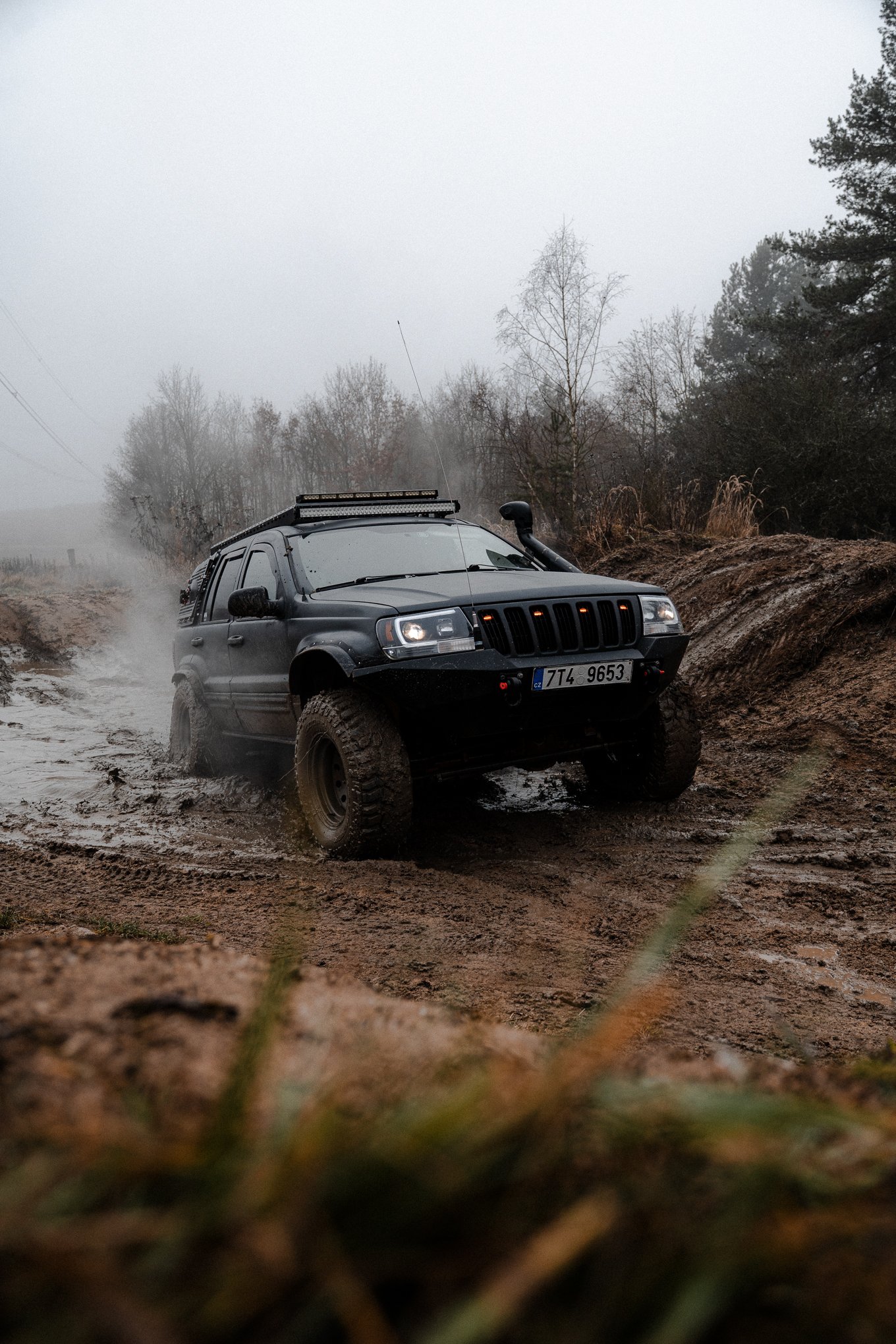 A black off-road vehicle driving through a muddy terrain on a foggy day, with trees in the background.