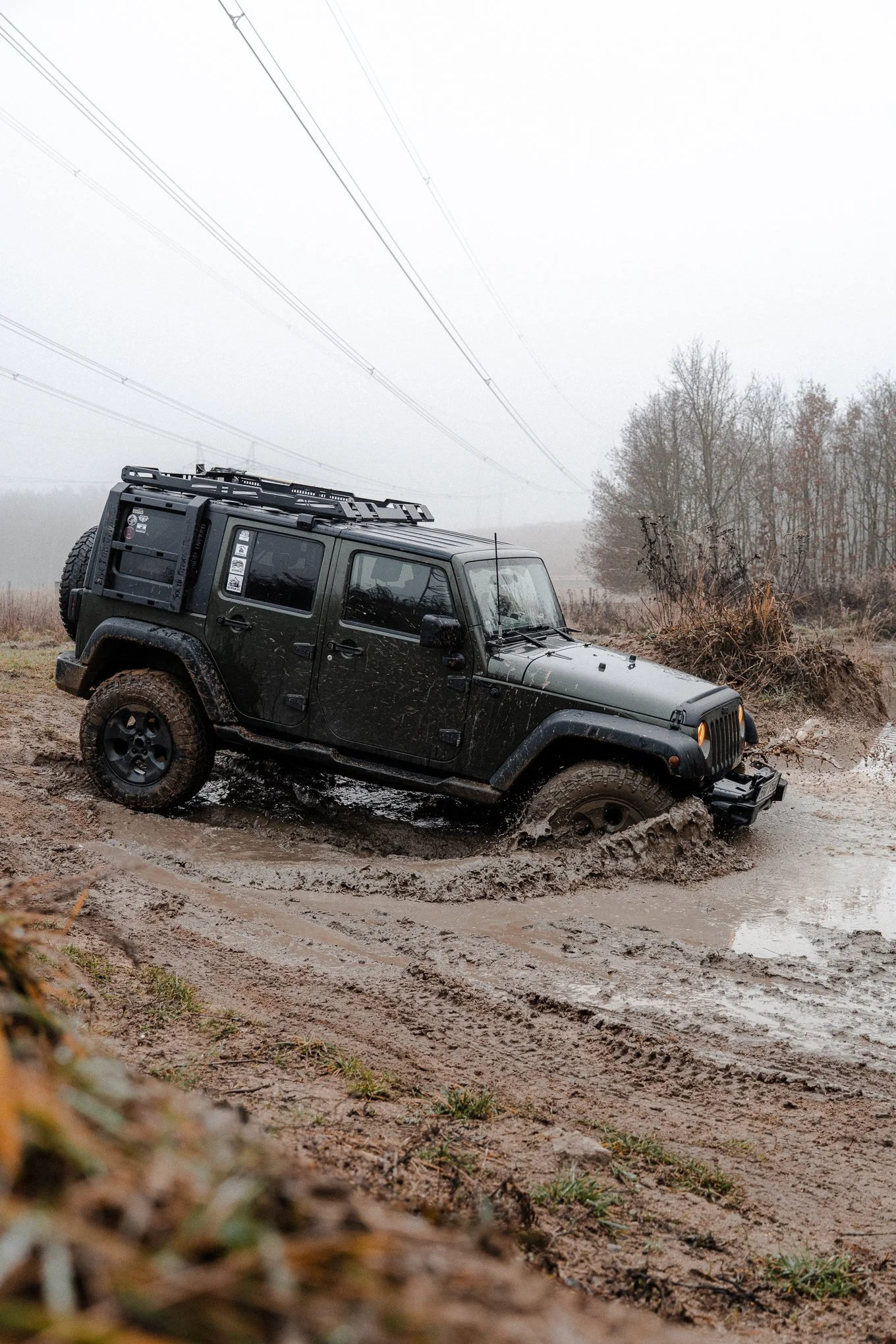 A black Jeep SUV driving through a muddy off-road trail in a rural area on a foggy day.