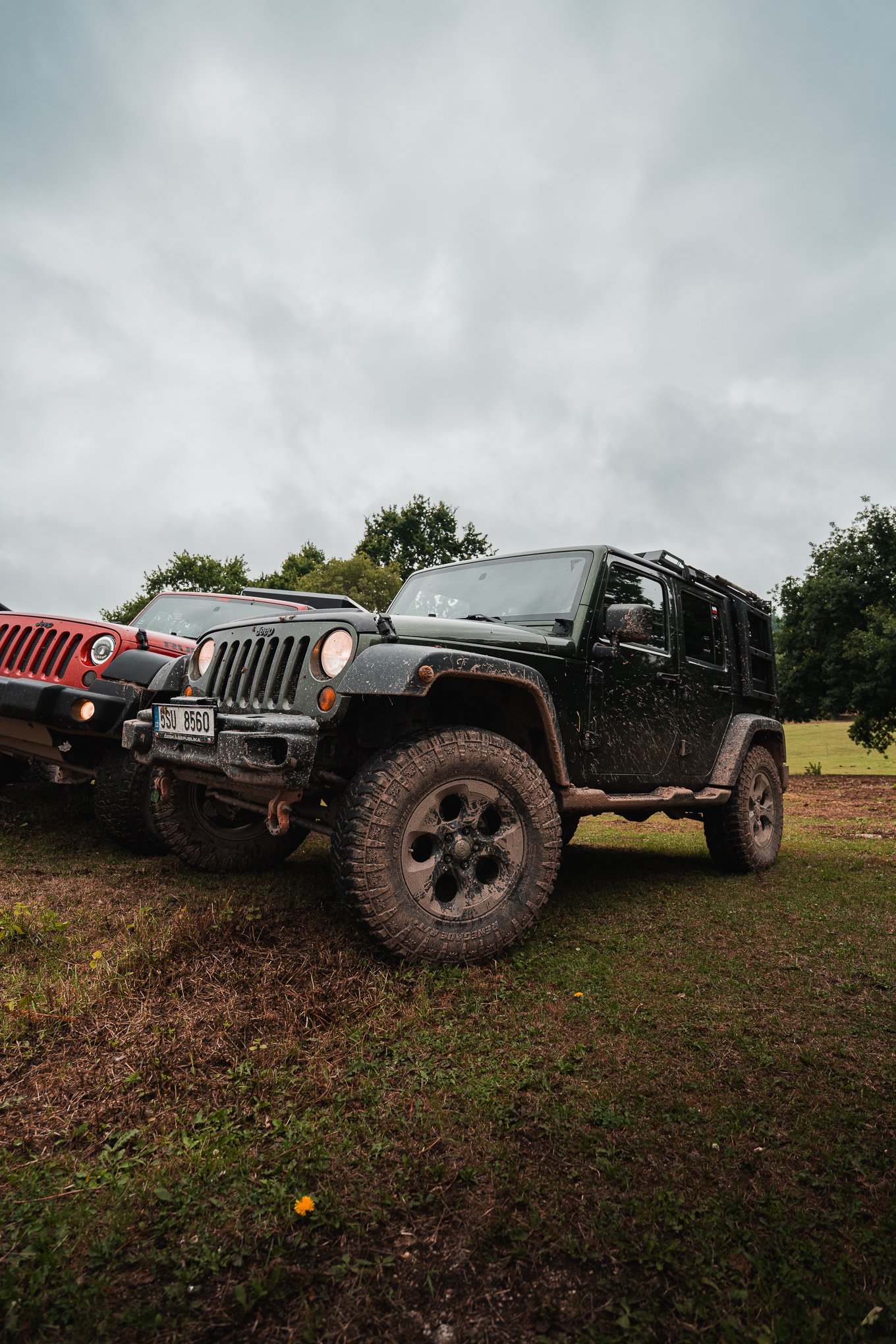 A black Jeep Wrangler with mud splatters parked on a grassy field under a cloudy sky.
