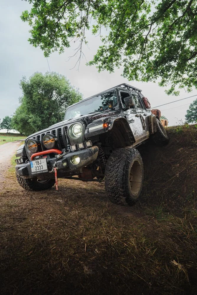 A four-wheel-drive off-road vehicle, likely a Jeep, tilted on a dirt embankment with one wheel in the air. It has large tires, a winch on the front bumper, and is surrounded by green trees and a cloudy sky.