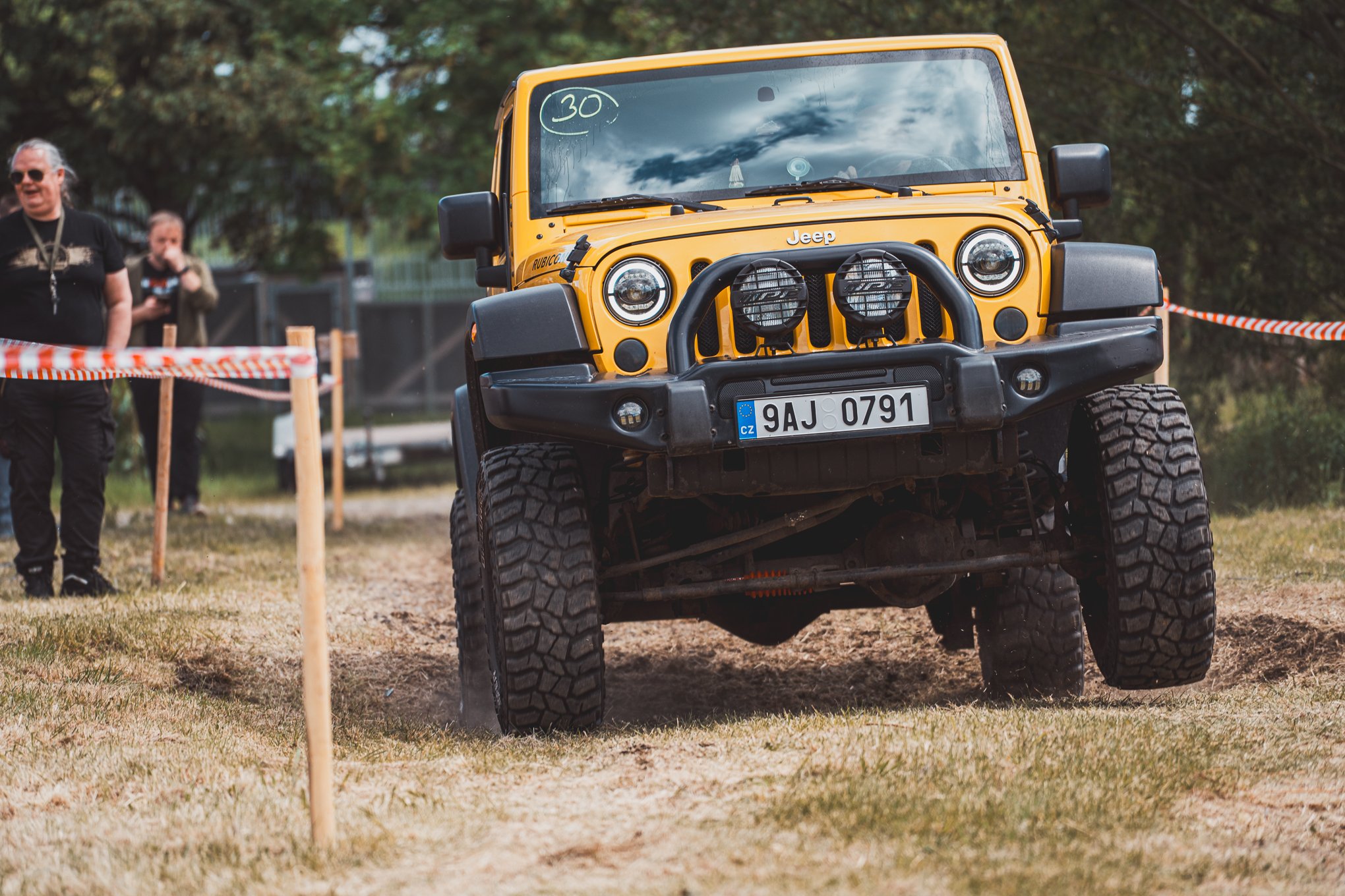 Yellow Jeep off-road vehicle driving on dirt path with front wheels lifted, surrounded by orange and white hazard tape, with people in black shirts watching in the background.