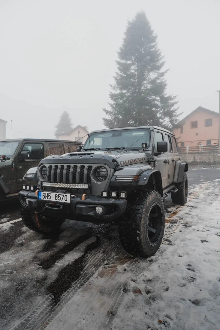 A gray Jeep Rubicon with off-road tires and bumper-mounted lights parked on a snowy, icy dirt road, with foggy trees and houses in the background.