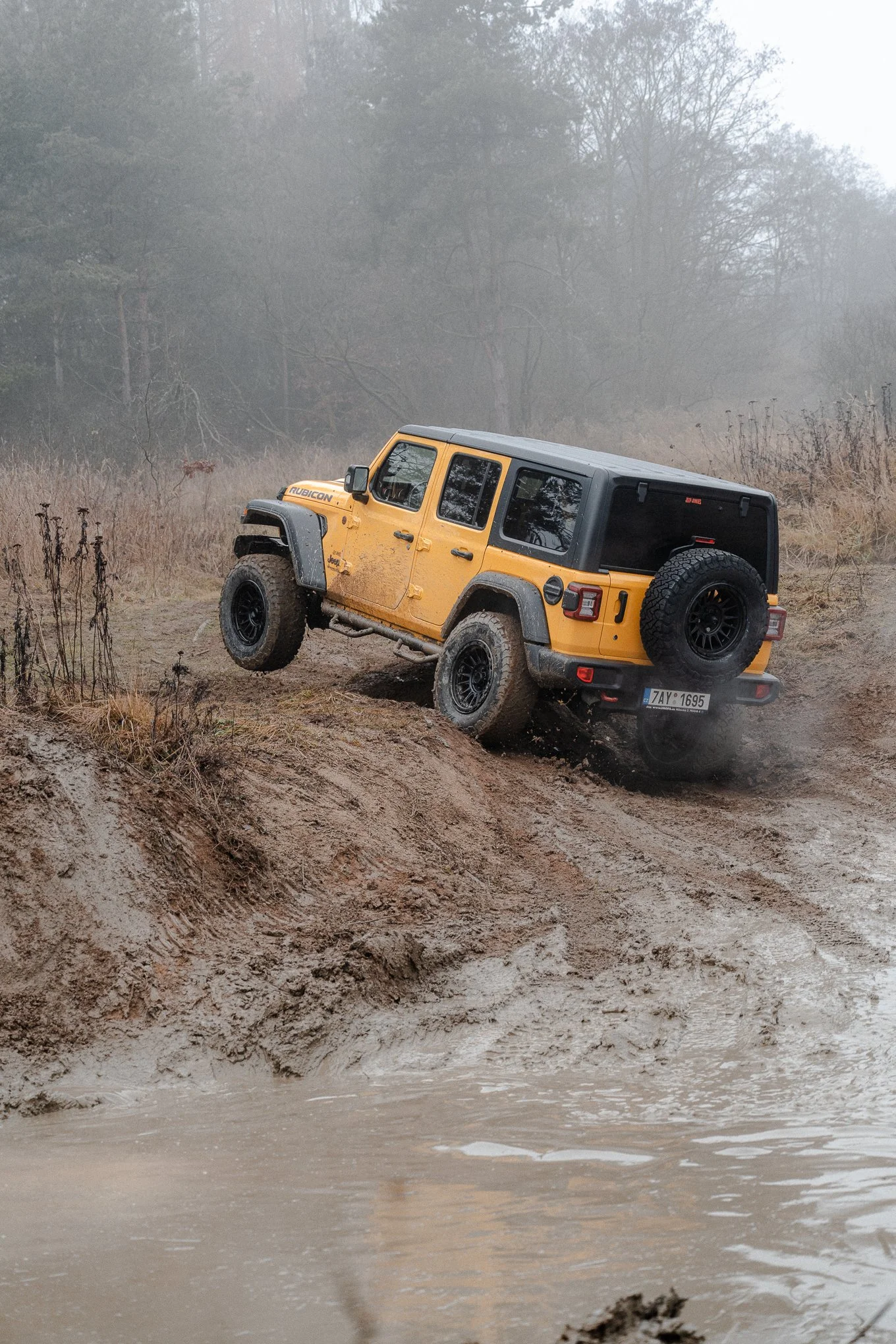 A yellow Jeep Wrangler Rubicon off-roading on a muddy terrain near a water body, with mud and dust being kicked up by the tires.