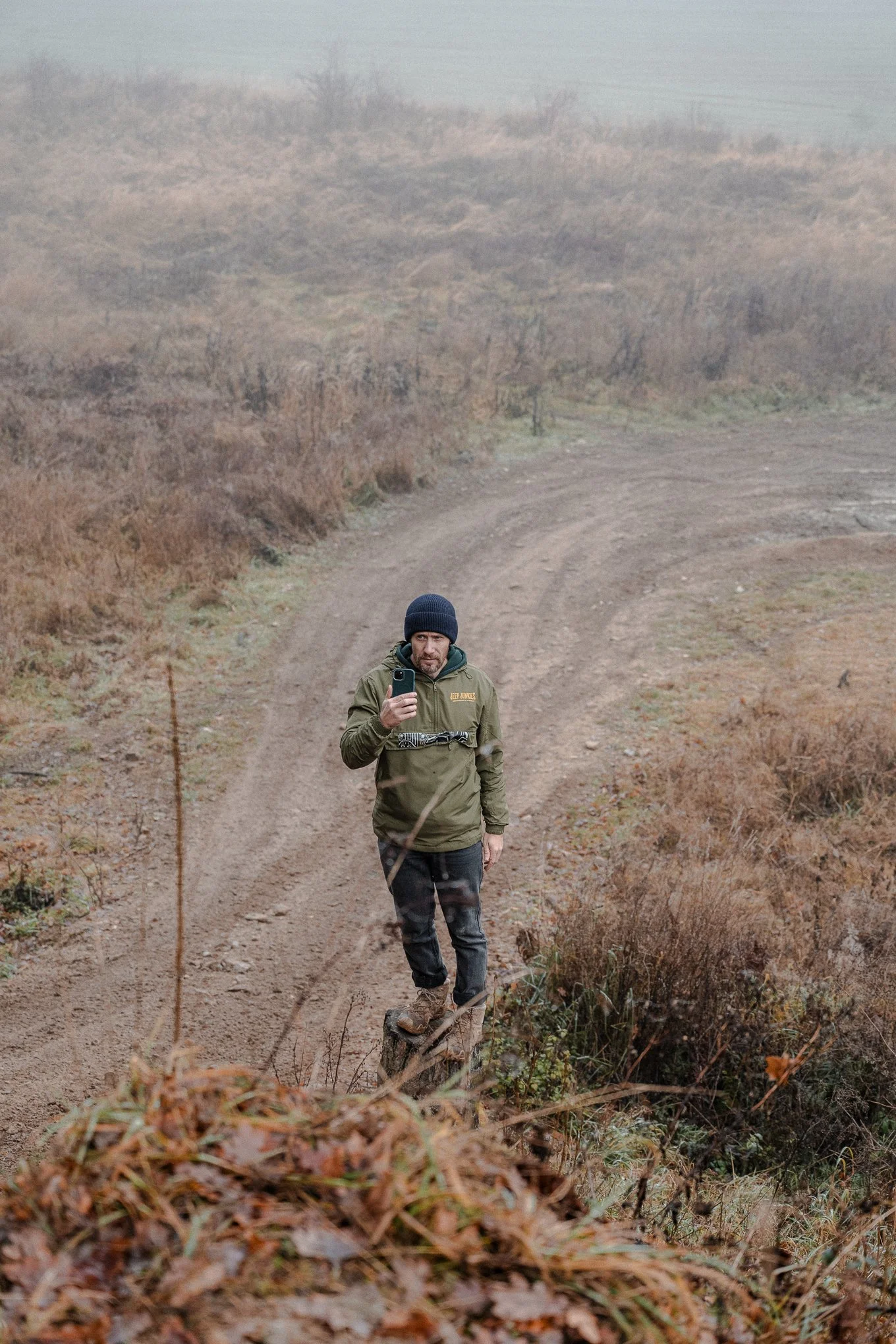 A man wearing a green jacket, black beanie, and hiking boots standing on a small tree stump taking a selfie along a dirt trail in a foggy, rural landscape with brown shrubs and barren trees.